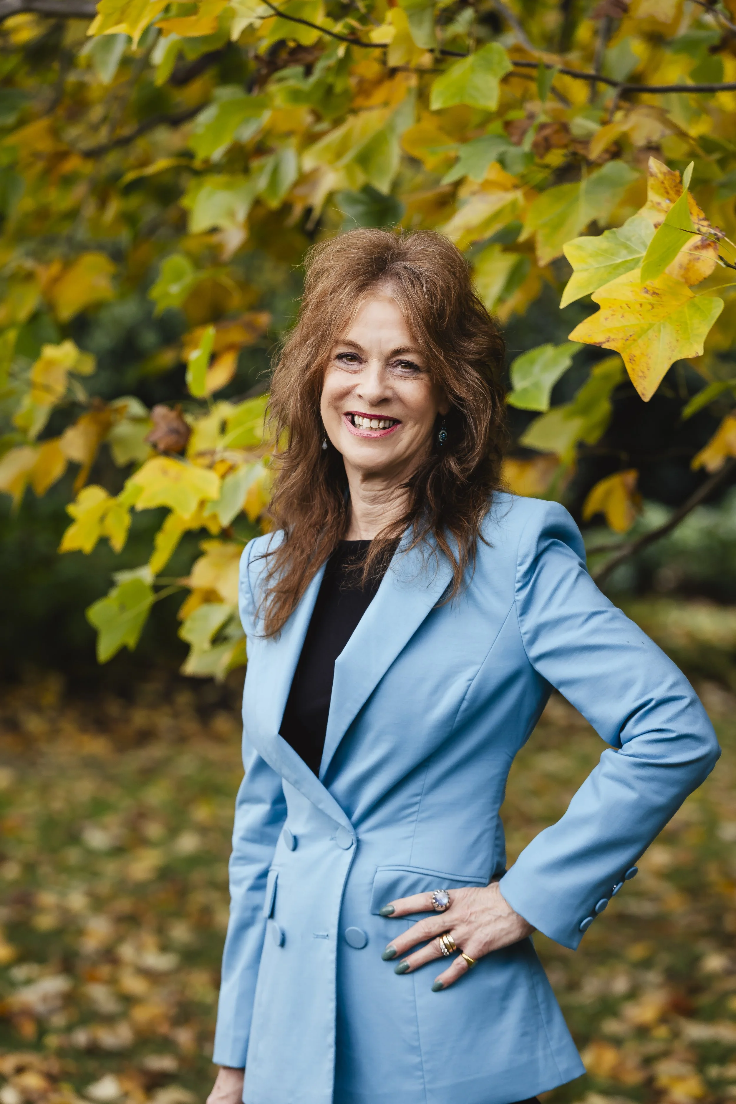 A woman with long brown hair smiling outdoors, wearing a light blue blazer over a black top, standing in front of yellow and green autumn leaves.