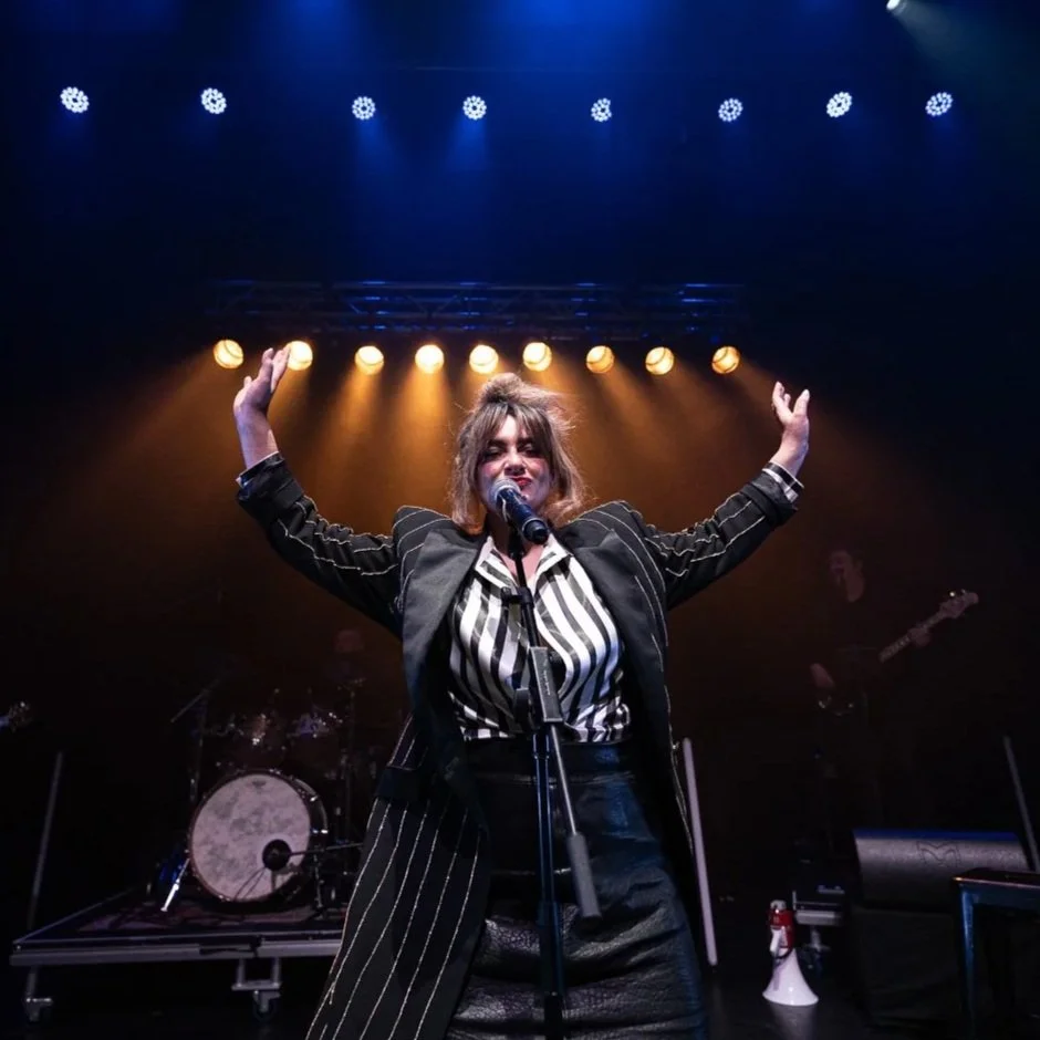 Performer on stage singing into a microphone with arms raised, band equipment visible in the background, stage lights shining overhead.