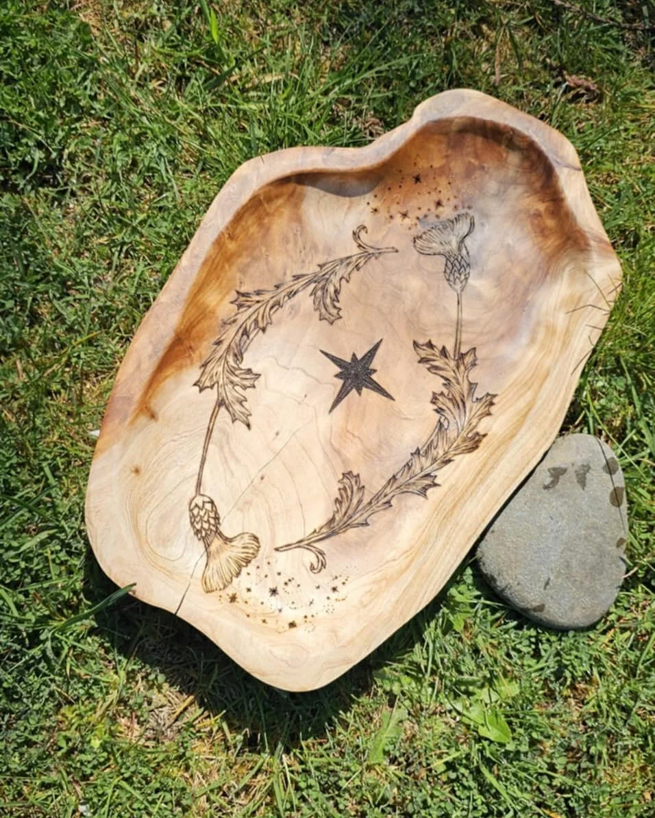 Wooden bowl with engraved floral and star patterns resting on grass with a gray stone nearby.