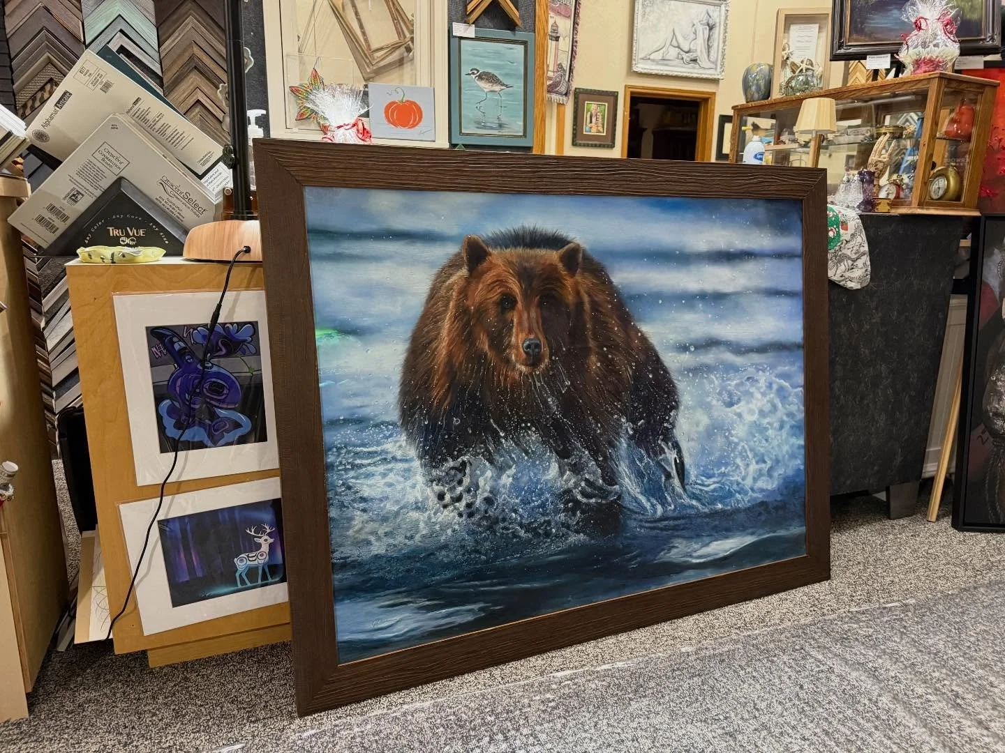 A framed painting of a brown bear walking in water with splashes, set in an art store with various other framed artwork and decorative objects in the background.