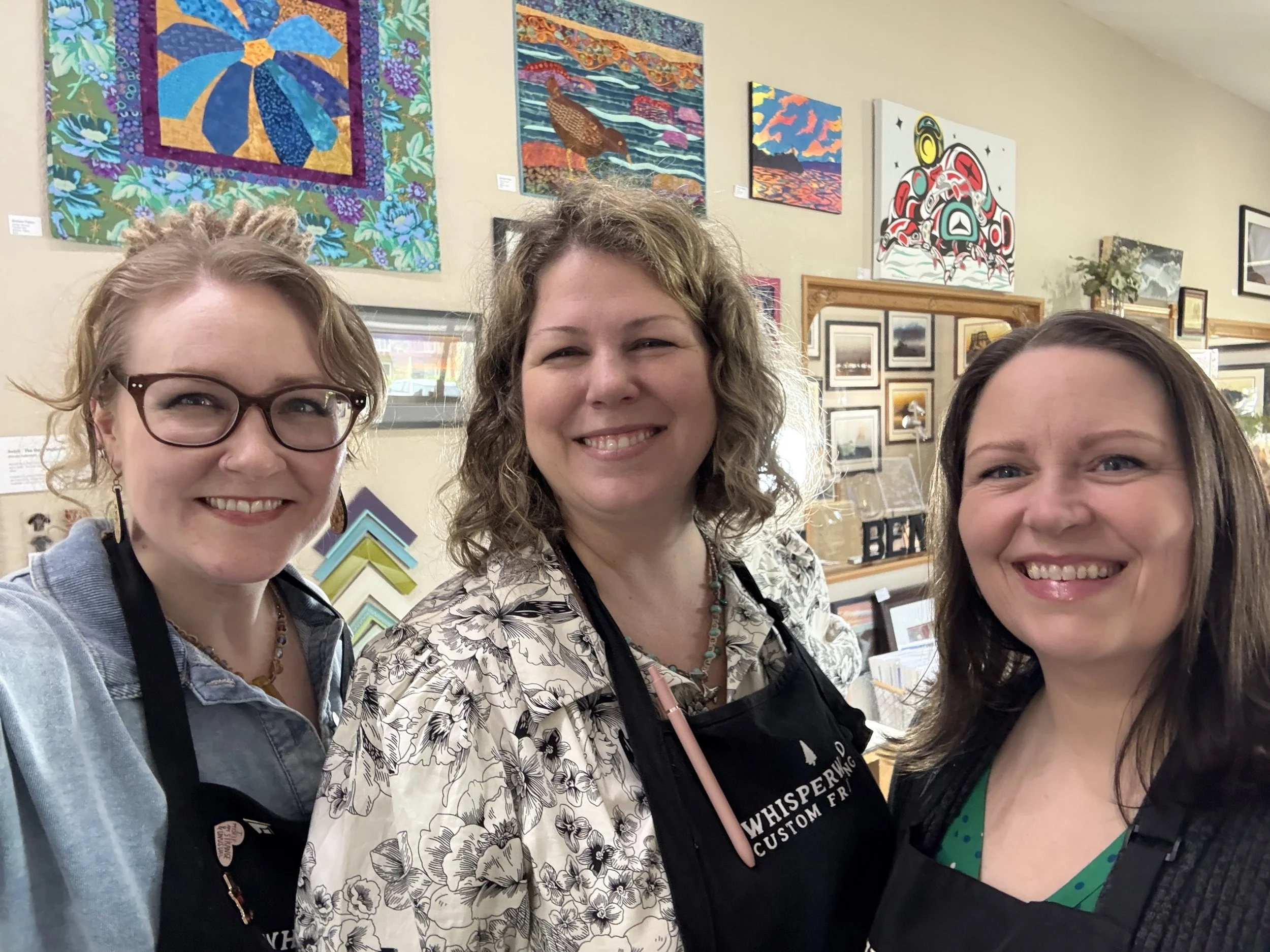 Three women smiling in an art gallery with colorful paintings on the wall behind them.