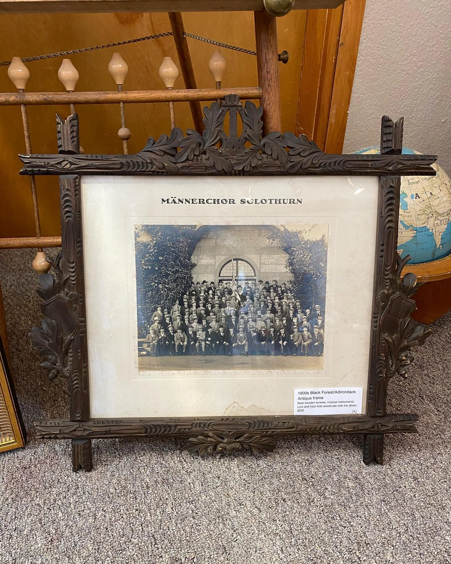 A vintage black forest/Adirondack picture frame made of carved wood with floral and musical instrument details. The frame contains a black and white photograph of a large group of people posing outdoors. There is a small white label on the bottom rig