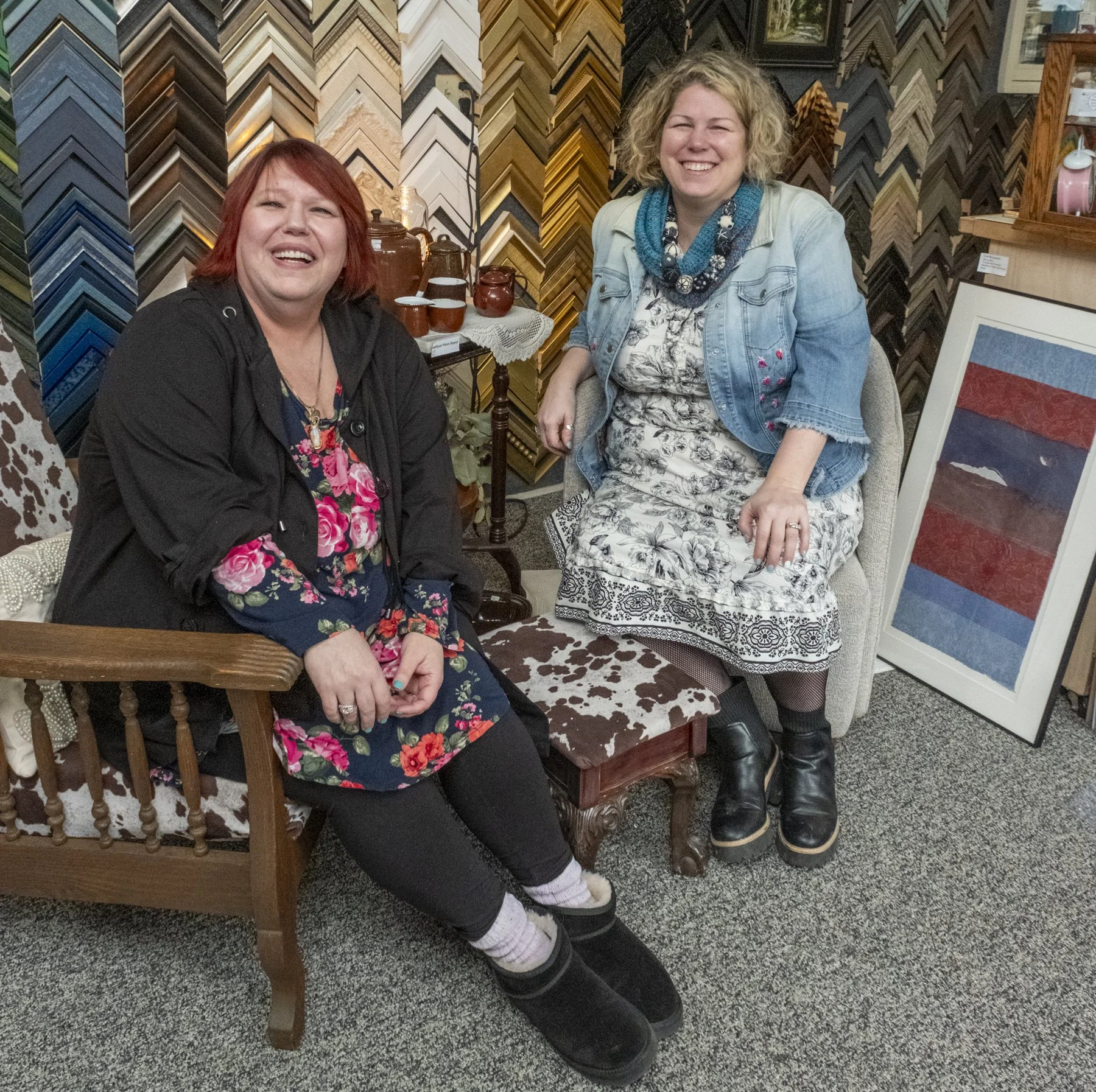 Two women sitting in a frame shop with colorful picture framing samples behind them, smiling at the camera, surrounded by various framed pictures and decorative items.
