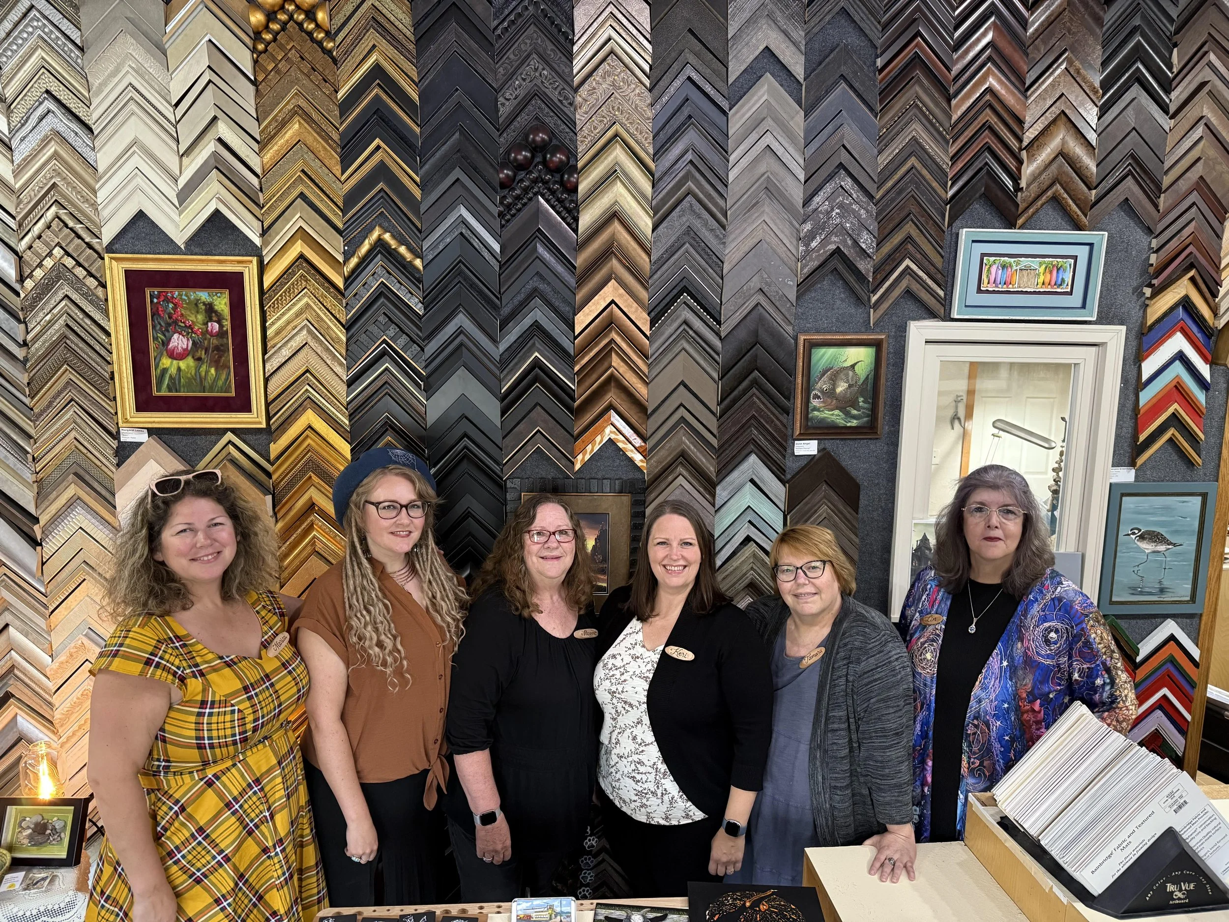 A group of seven women standing inside a picture framing shop with various picture frames and artwork displayed on the wall behind them.