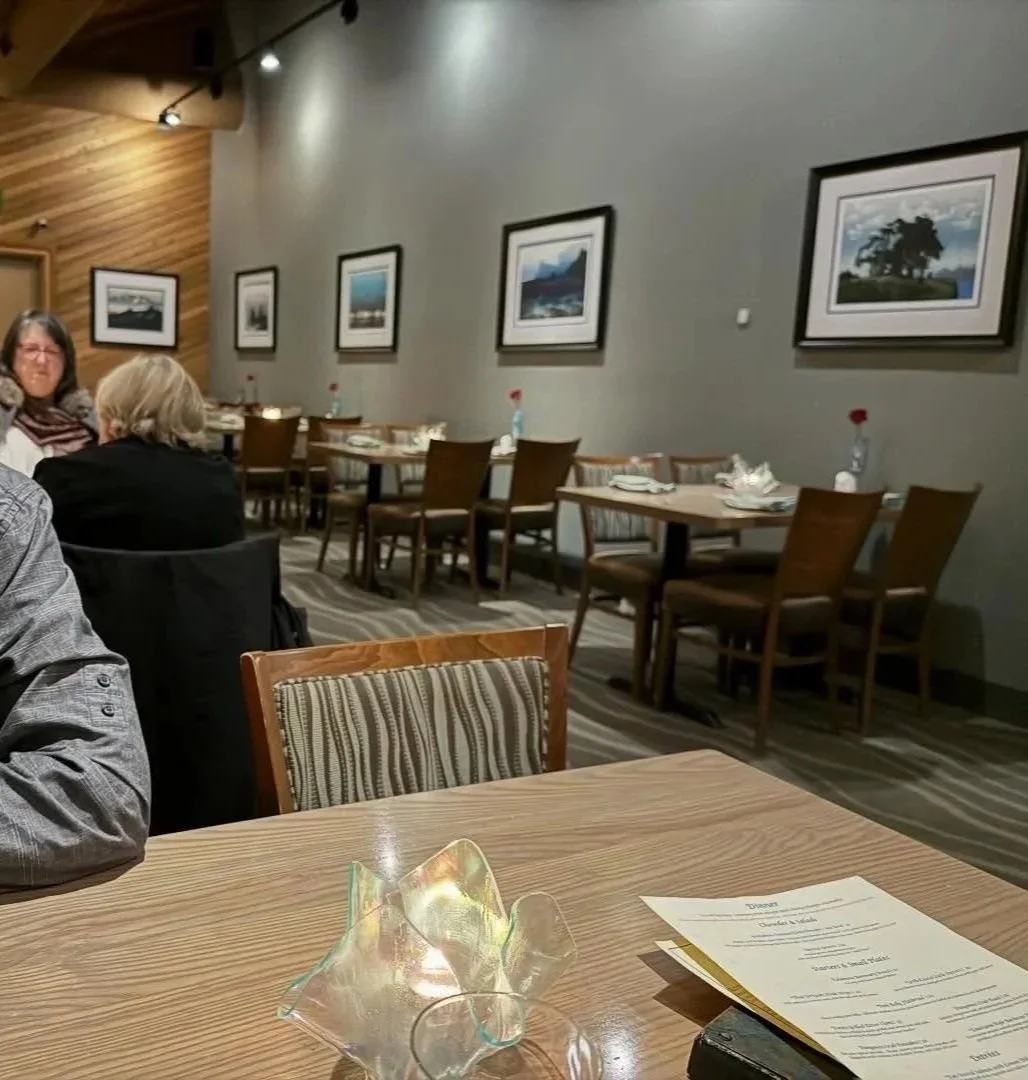 Inside a restaurant with wooden and gray walls, framed landscape photos, tables set with napkins, water bottles, and utensils, some guests seated, a colorful glass decoration on a wooden table in the foreground.