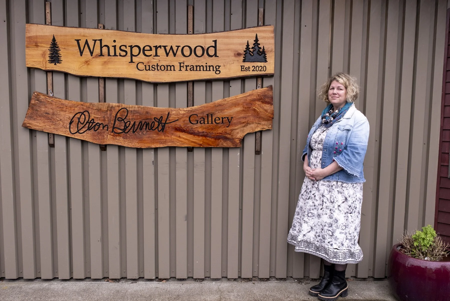 A woman standing beside a large wooden sign on a gray corrugated metal wall. The sign reads "Whisperwood Custom Framing" with a tree graphic and 
