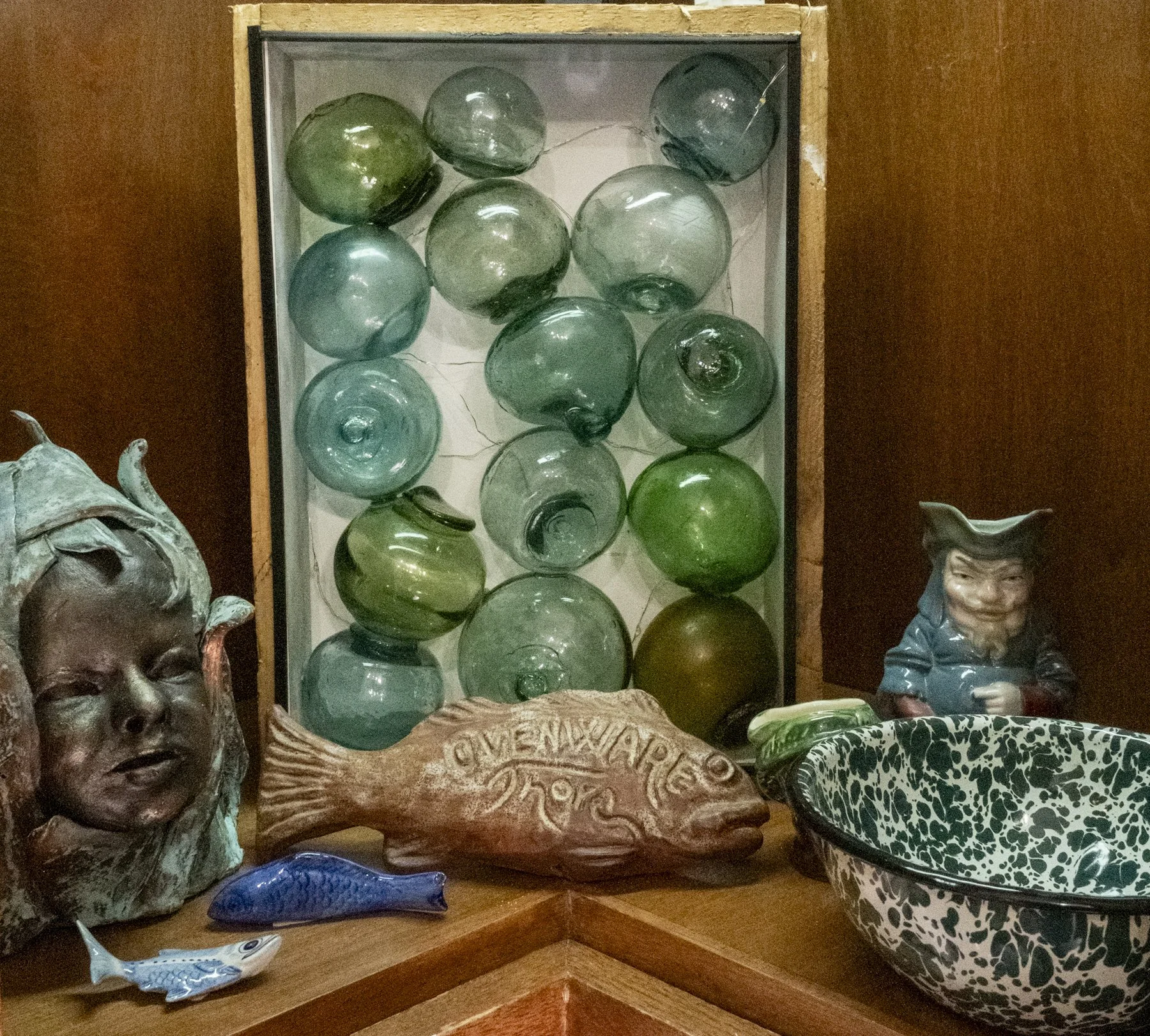 Display of decorative glass balls in varying shades of green on a shelf, flanked by ceramic figurines of faces, with a patterned bowl and a carved wooden sign reading "ONWARD" in the foreground.