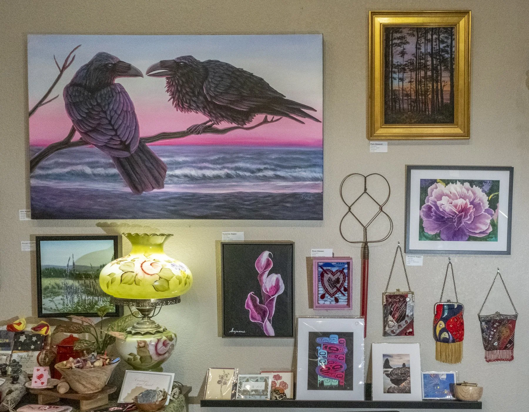 Art gallery wall with paintings including a fox and eagle on a beach at sunset, a forest scene in a golden frame, and a flower. Decorative items include a floral lamp, jewelry, and small framed artworks.