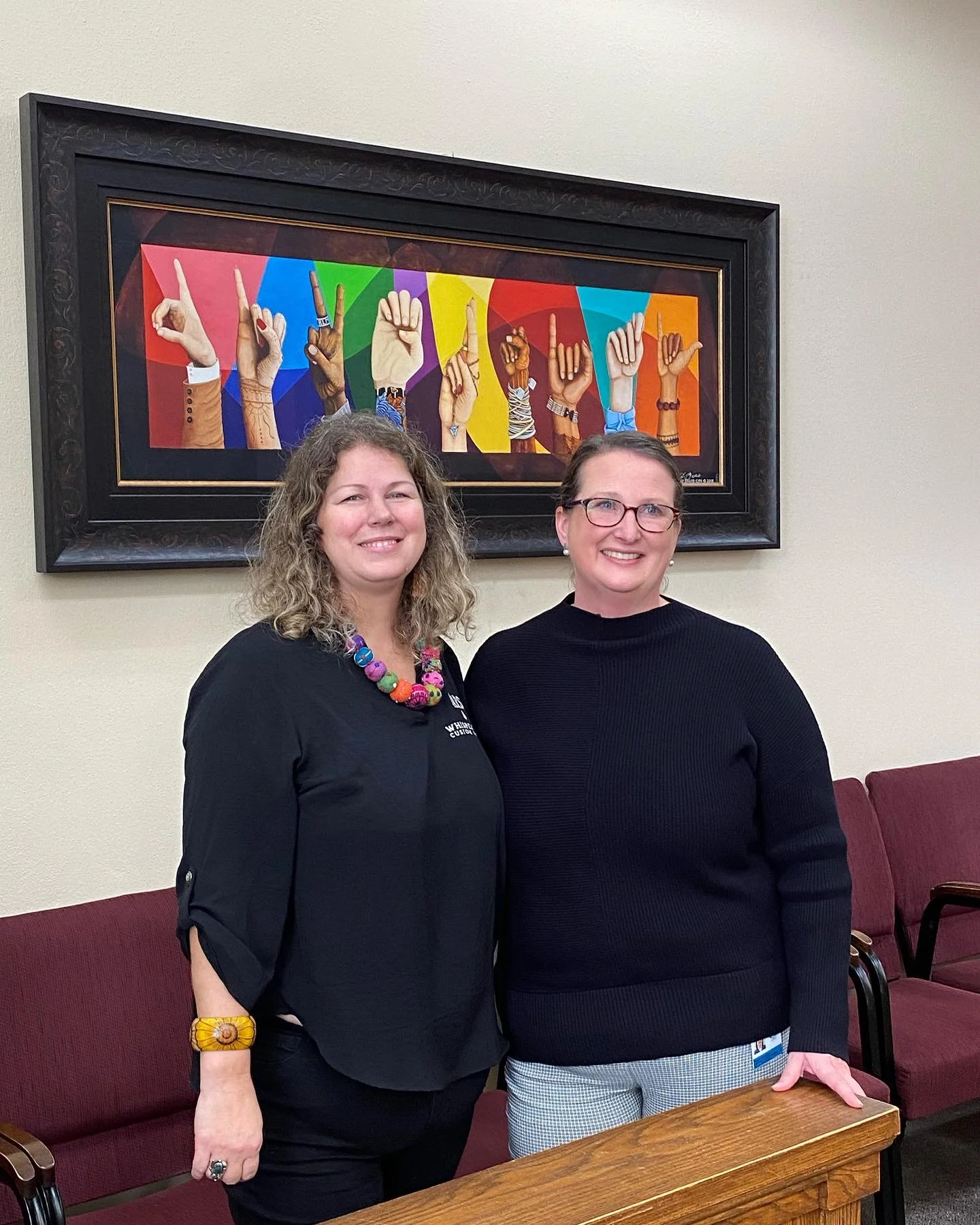 Two women standing inside a room with a framed colorful artwork of diverse hands making various gestures in the background. They are smiling and dressed in black clothing, with chairs and a wooden table visible in the room.