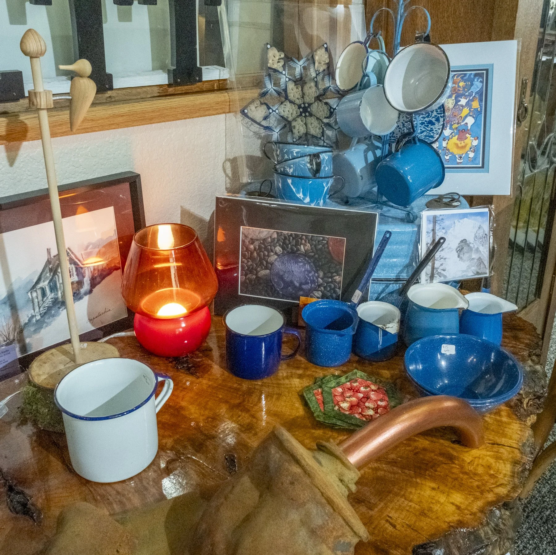 Display of blue and white enamel mugs, bowls, and cups on a wooden table, along with framed artwork and a red glass lamp.