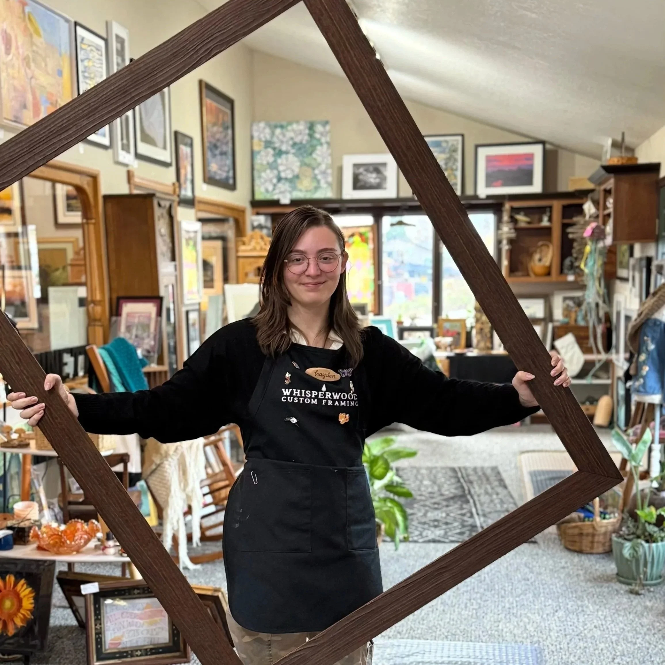 A woman with glasses and long dark hair smiling inside an art or gift shop, holding a large wooden picture frame in front of her. The background shows various framed pictures and decorative items displayed on shelves and walls.