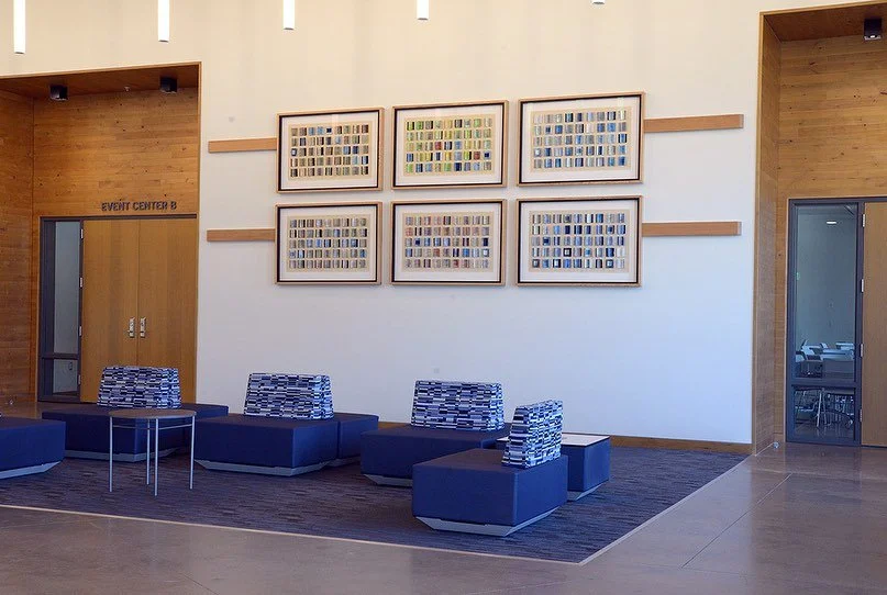 Waiting area with six blue and patterned chairs, artwork of framed colorful contacts on the wall, wooden wall panels, and a door labeled 'EVENT CENTER B.'