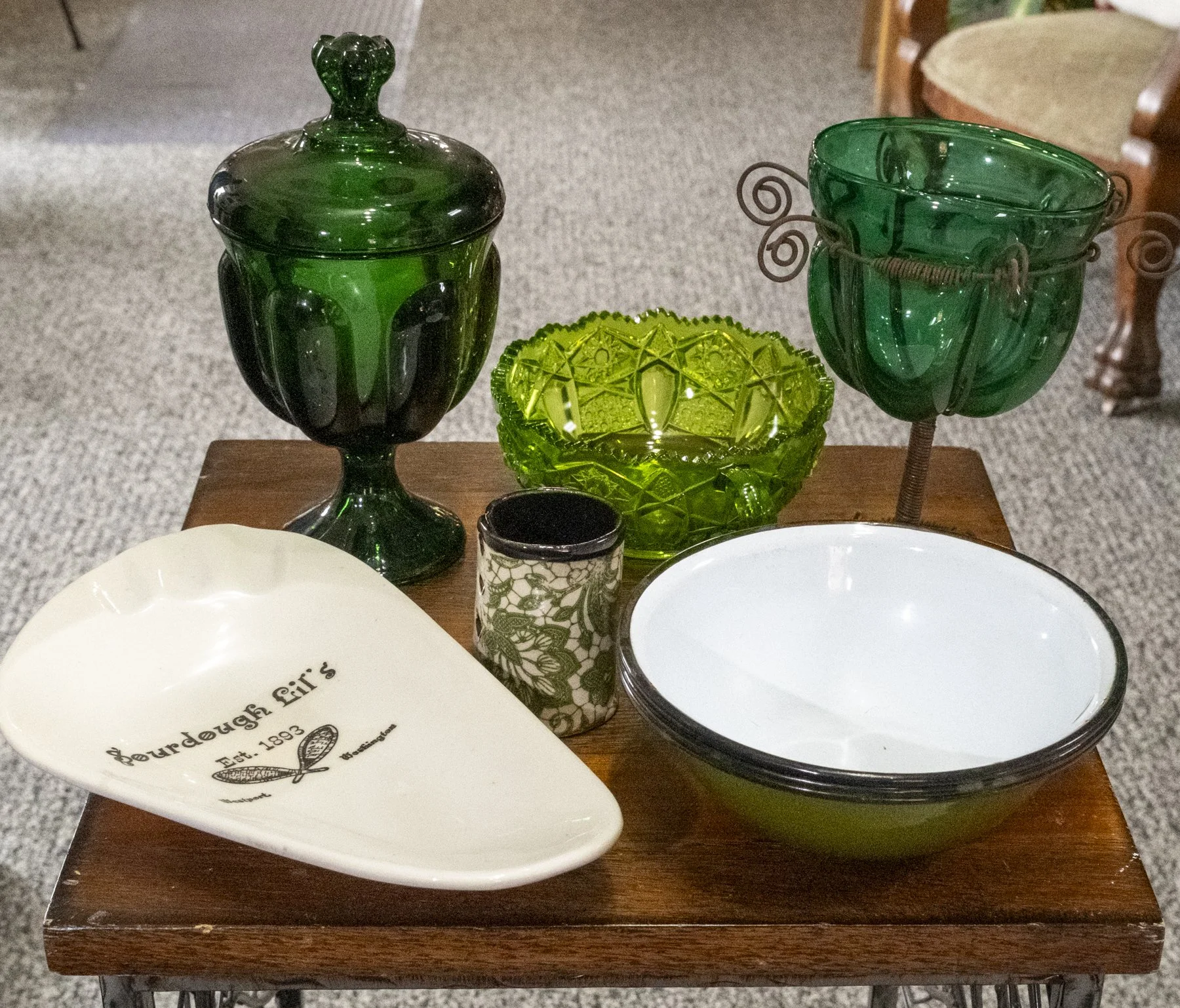Green glass jar with a lid, a red and green glass bowl, a small green bowl, a black and white decorative cup, a large green and black bowl, and a white oval dish on a wooden surface.