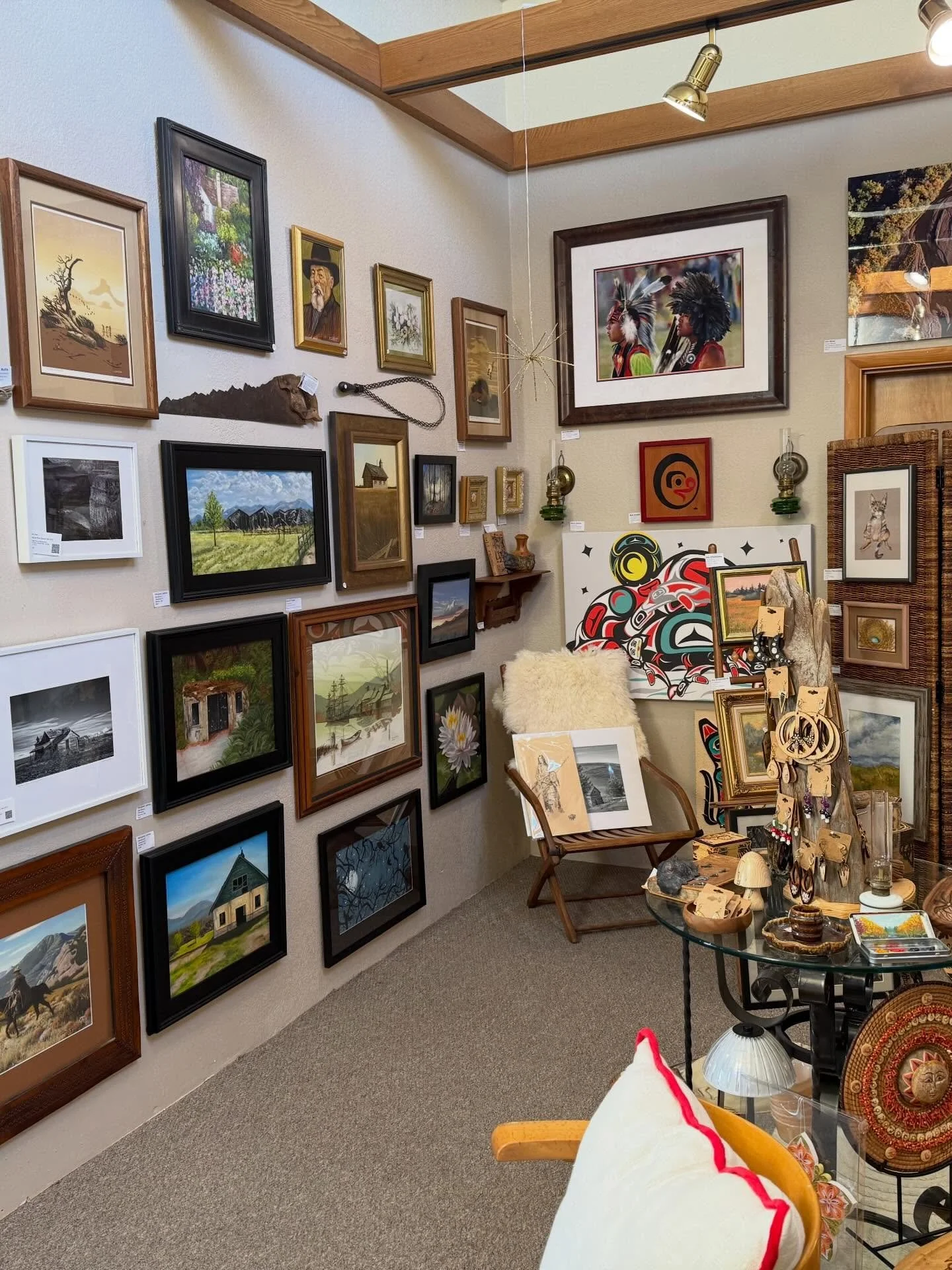 An art gallery displaying various paintings and artwork, with a small table holding decorative items, and a chair with a sheepskin cushion.