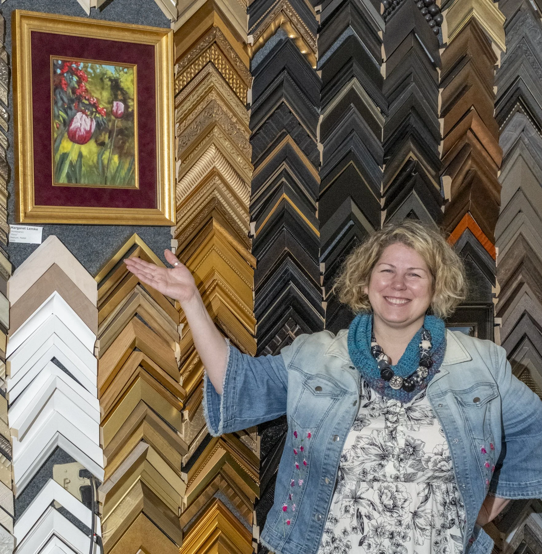 A woman with curly blonde hair smiling at a picture framing store, standing in front of a large display of various picture frame samples. She is wearing a floral dress, a light denim jacket, and a blue beaded necklace. She is gesturing towards the fr