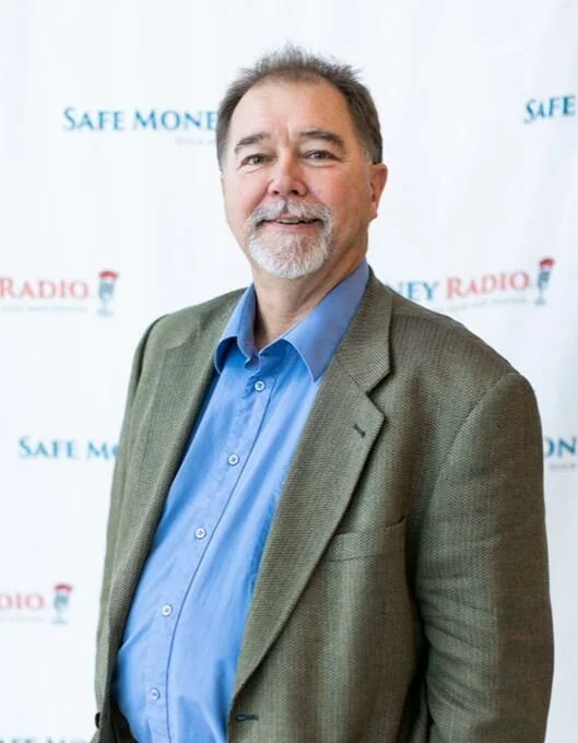 A middle-aged man with gray hair and a goatee, wearing a blue dress shirt and a gray blazer, standing in front of a backdrop with logos for Safe Money and Radio.