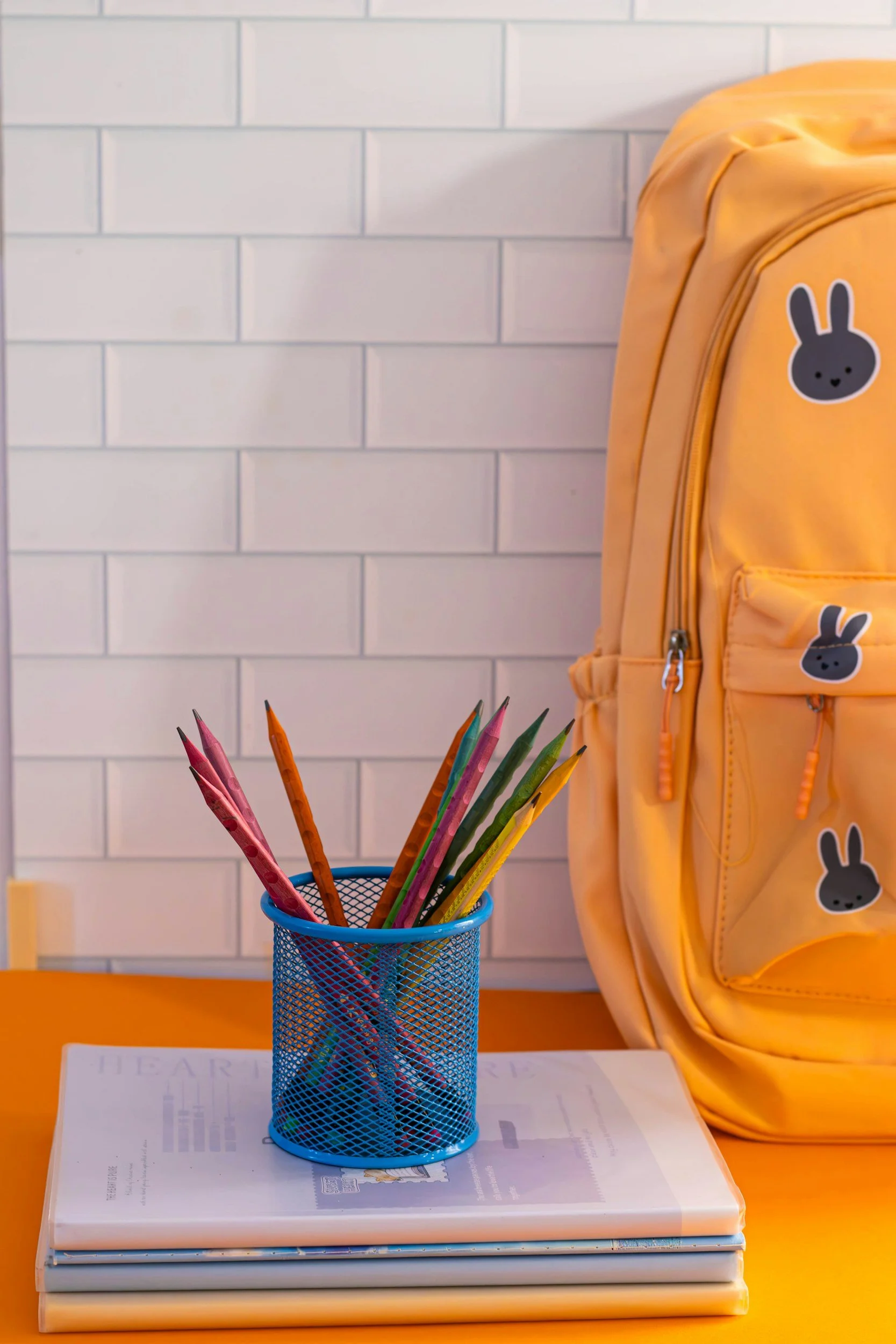 A yellow backpack with bunny face stickers on a white-tiled wall, a blue pencil holder with colorful pencils, on top of a closed book or magazine on an orange desk.