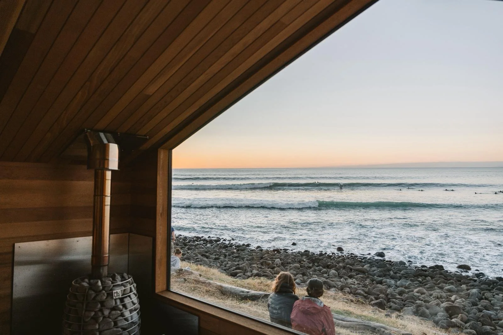 View of the ocean through a large window from inside the Yutori Sauna, with people sitting on rocks near the shoreline during sunset.
