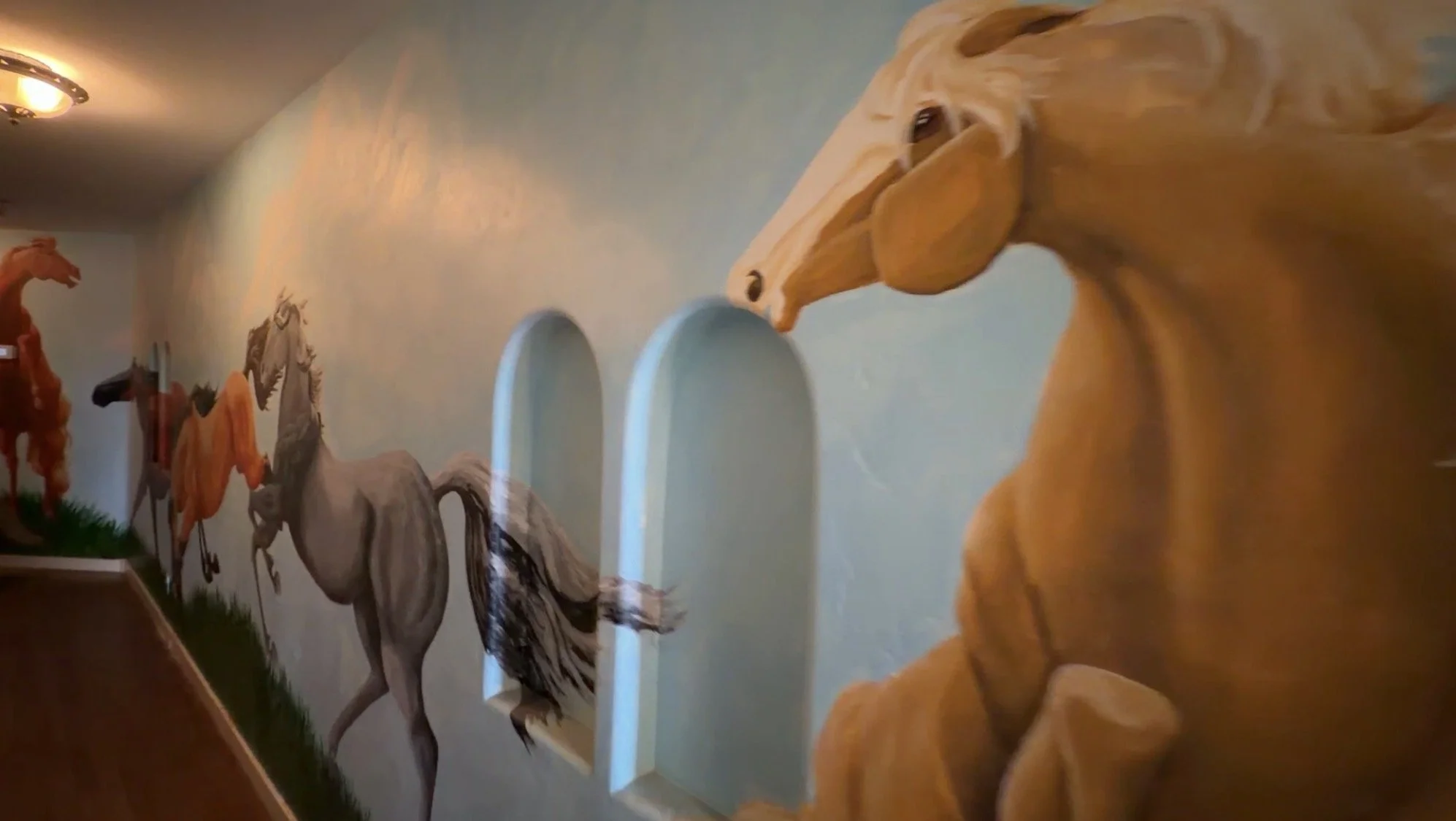 Mural of painted horses in various colors, including orange, gray, and black, on a hallway wall with arched doorways.