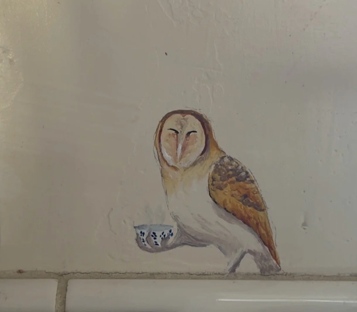 Hand-painted artwork of an owl with closed eyes, holding a small blue and white patterned bowl, on a white wall.