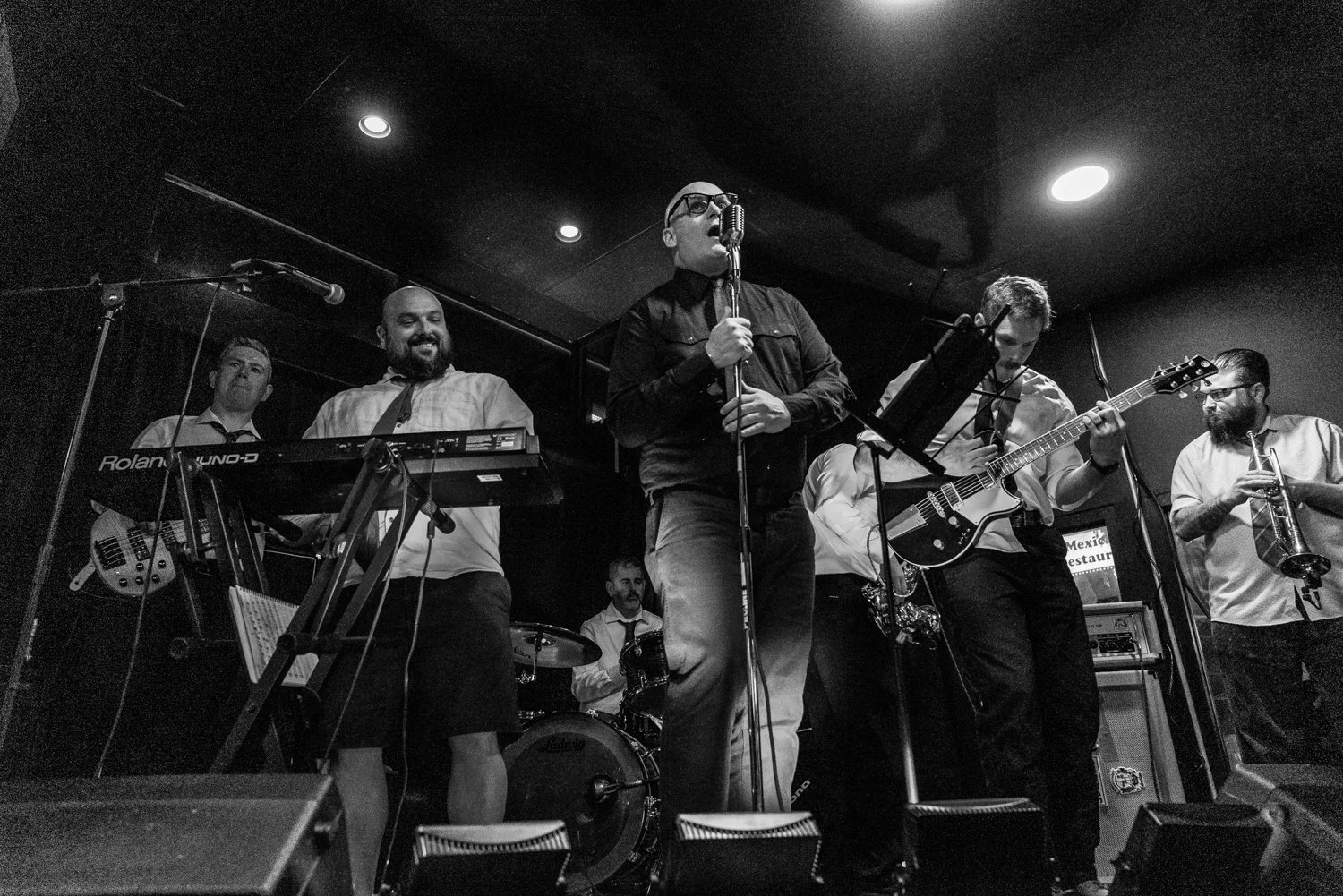 A black and white photo of a live band performing on stage. The band has five members: a keyboard player, a singer with glasses holding a microphone, a drummer, a guitarist, and a saxophonist. They are in a dimly lit venue.