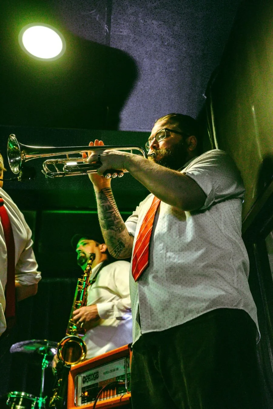 Man with glasses playing trumpet in a band, with a saxophonist in the background and a green-lit stage.