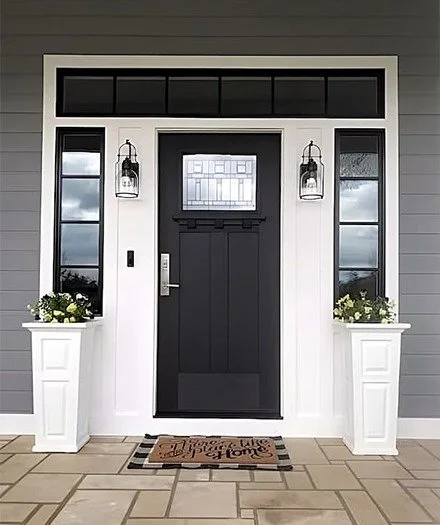 Front door of a house with a black door, two large planters with flowers, two wall-mounted lantern lights, and transom windows.