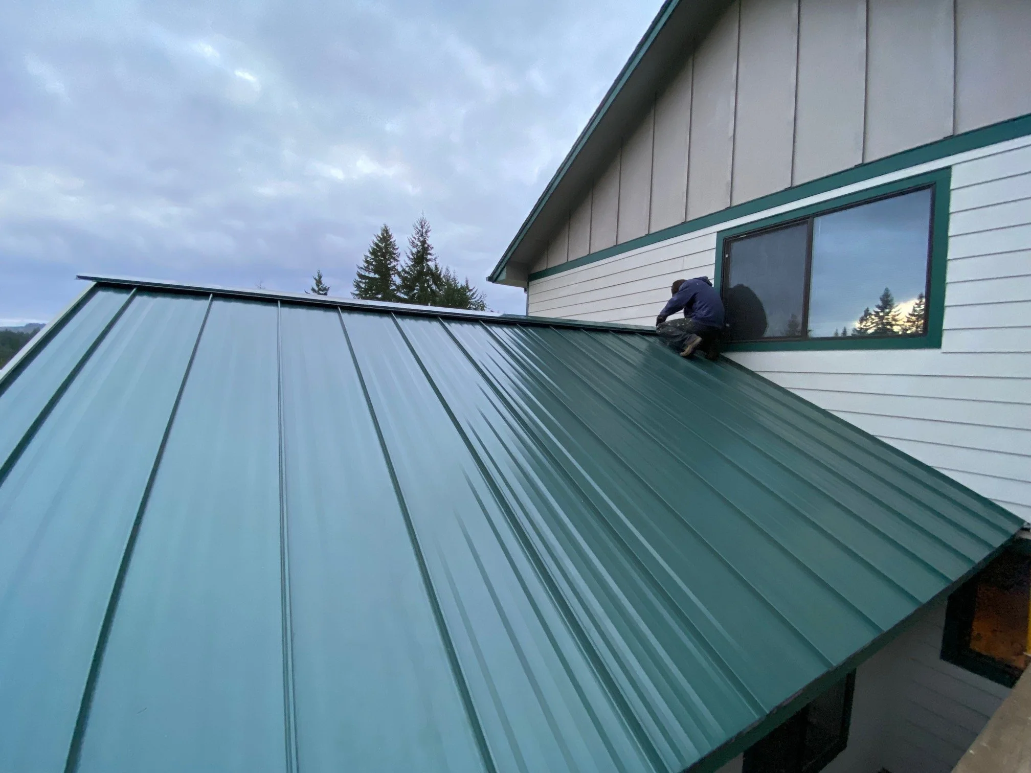 A person installing a dark green metal roof on a house with white siding and a large window, trees in the background, during cloudy weather.