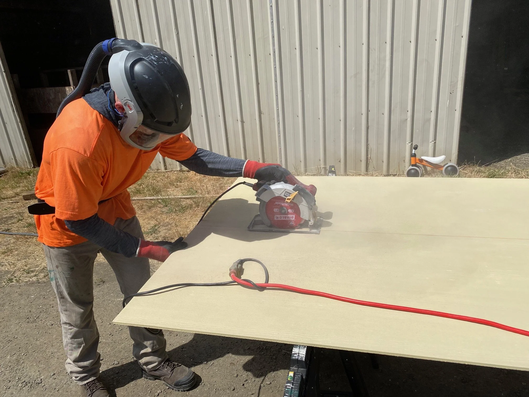 A person wearing safety gear cuts a large sheet of wood with a power saw outdoors, with a metal building and a small bicycle in the background.