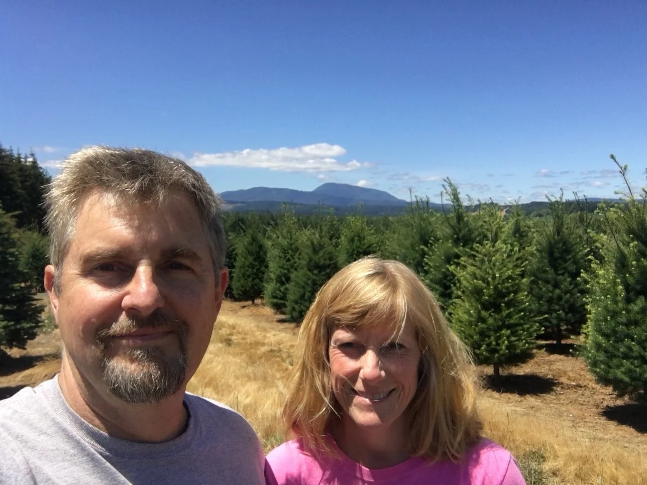A man and woman smiling outdoors in a Christmas tree farm with rows of evergreen trees and mountains in the background under a blue sky with clouds.