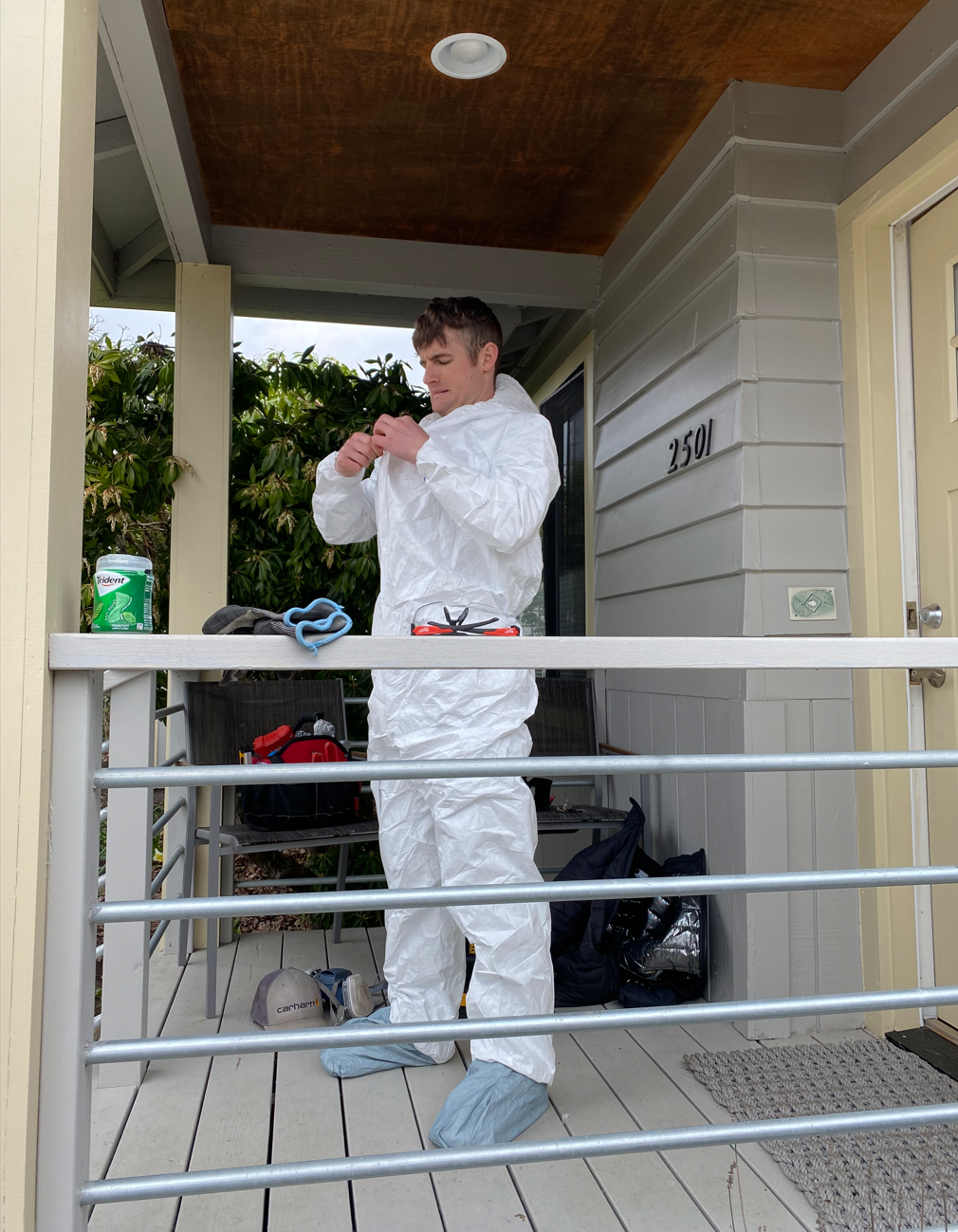 A man in white protective coveralls and shoe covers stands on a porch, preparing for work. He is surrounded by tools, a black bag, and a green container of disinfectant wipes, with a background of green bushes.