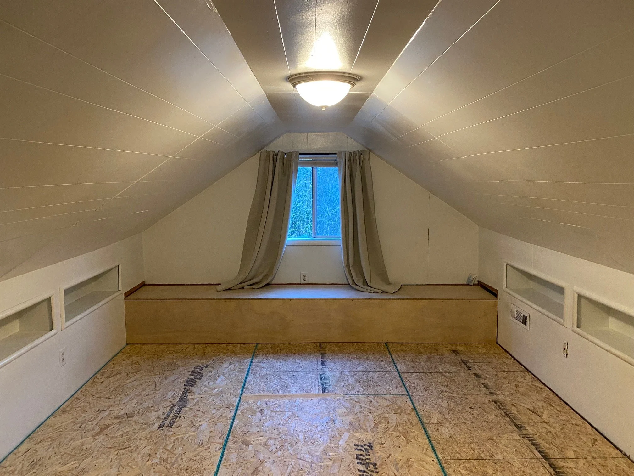 Empty attic room with sloped ceiling, window with curtains, and unfinished flooring.