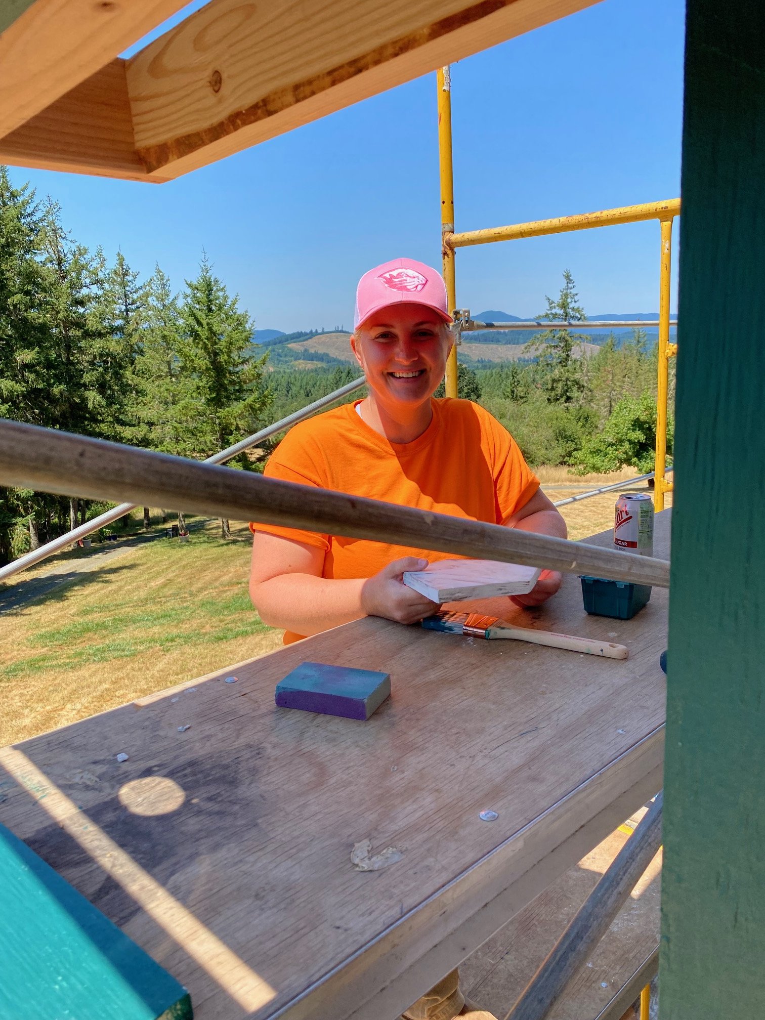A woman wearing an orange shirt and pink cap smiling and working on a construction project at a scenic outdoor location. She is holding a paintbrush and sitting at a wooden work surface with construction tools on it, with a backdrop of trees, hills, and a clear blue sky.