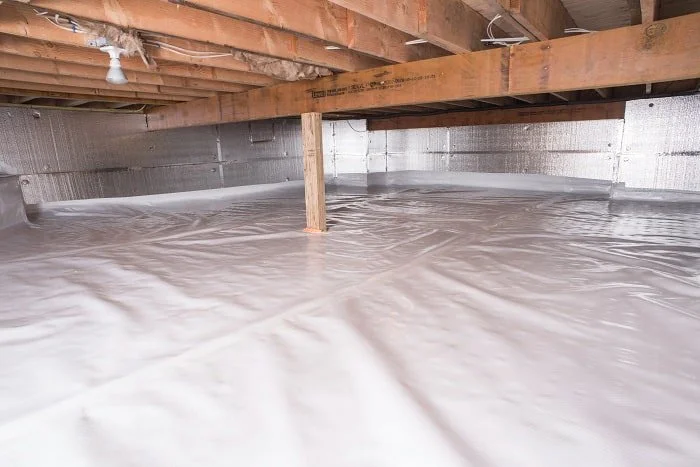 A crawl space with a plastic vapor barrier installed on the ground and wooden beams supporting the floor above.