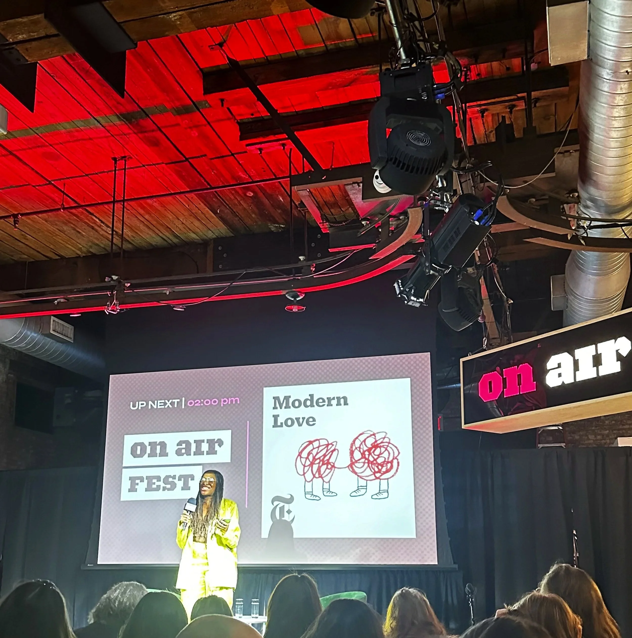 A woman in yellow clothing stands on stage at a conference with a large screen behind her. The screen displays the event name 'on air FEST', the upcoming segment titled 'Modern Love' at 2:00 pm, and an artistic drawing of two lions with tangled manes. The audience is visible in the foreground, and the stage area has black curtains. Overhead, stage lighting and audio equipment hang from the ceiling.