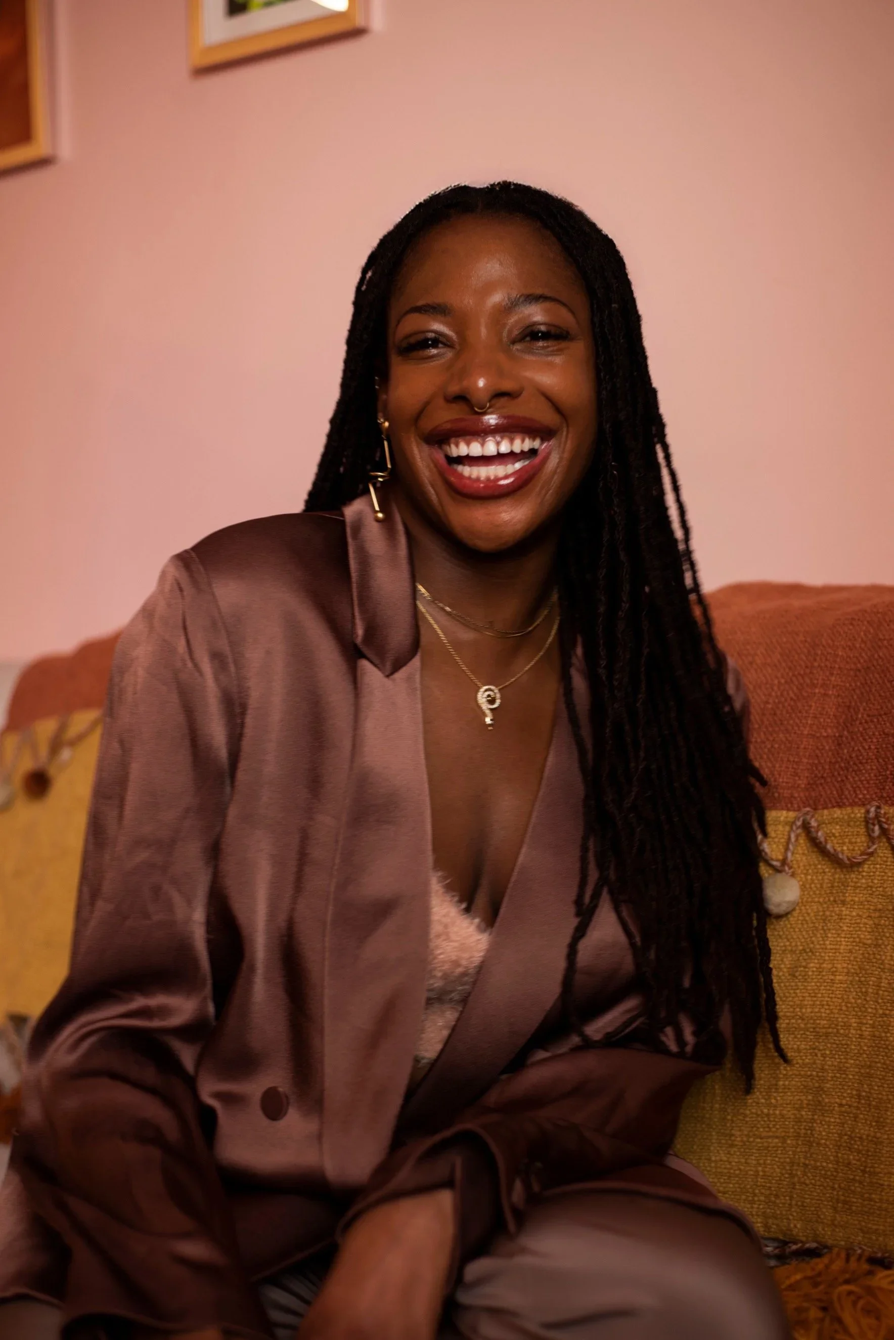 A woman with long dreadlocks smiling, wearing a satin blazer and jewelry, sitting on a sofa with a pink wall and framed pictures in the background.