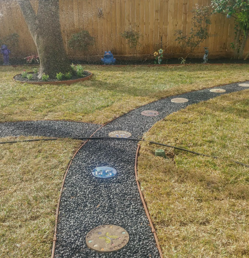 A backyard with a curved pathway made of black gravel and colorful stepping stones. There is a wooden fence at the back, a large tree with a flower bed around its base, and decorative garden statues along the fence.