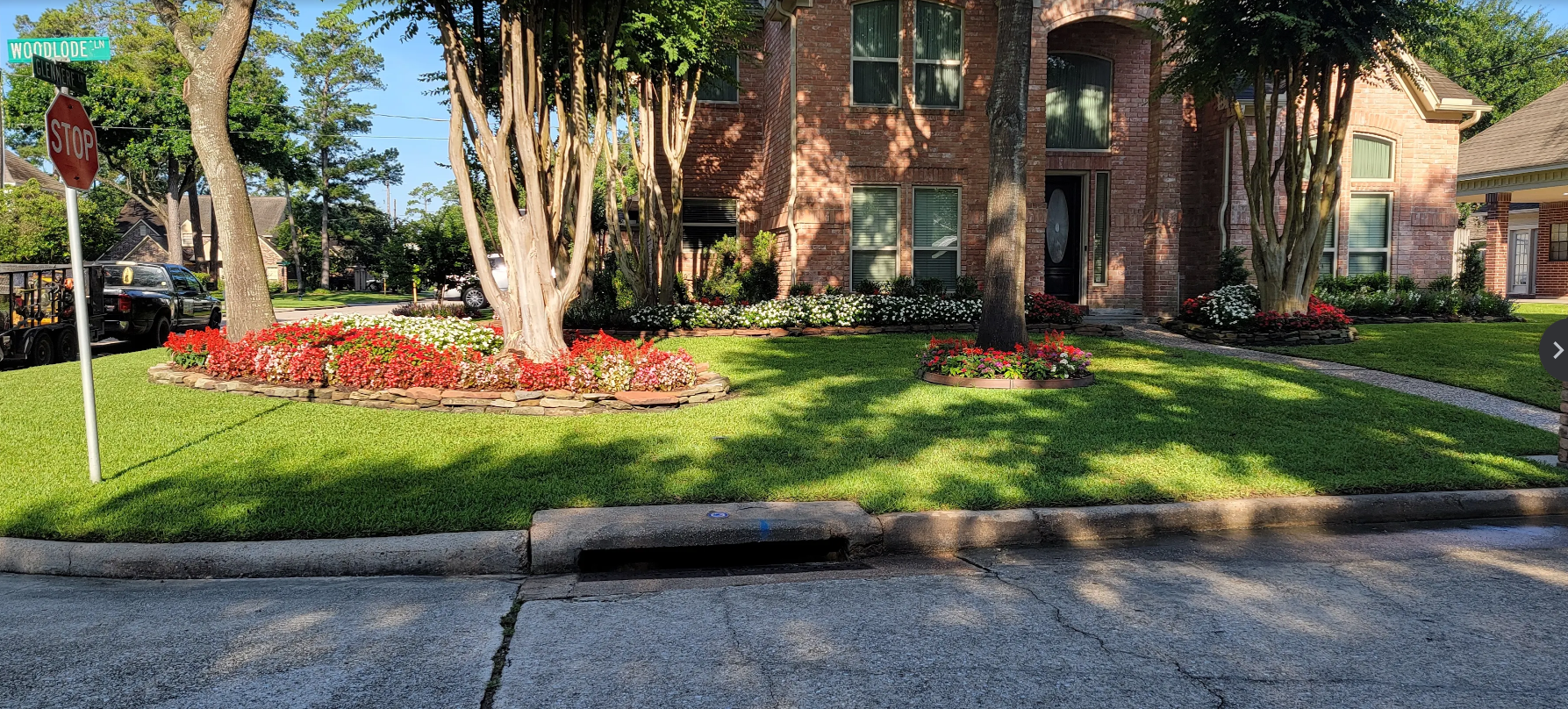 A well-maintained front yard of a brick house featuring lush green grass, colorful flower beds with red, white, and pink flowers, and mature trees with trimmed trunks. There are street signs at the corner of the yard, one indicating a stop sign and the other showing street names 'Woodlode Ln' and 'Glenmere Ln'. The driveway is visible at the front, and part of a neighboring house is seen to the right.