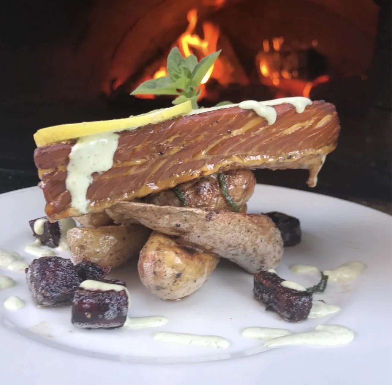 A plated dish with grilled salmon, roasted potatoes, baked beets, a slice of lemon,  and a garnish of herbs, with a background of a fire in a pizza oven.
