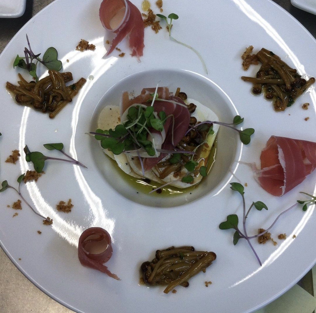 An elegant plate of gourmet food featuring thinly sliced cured meats, microgreens, and caramelized mushrooms arranged artistically with drizzles of oil and garnishes.