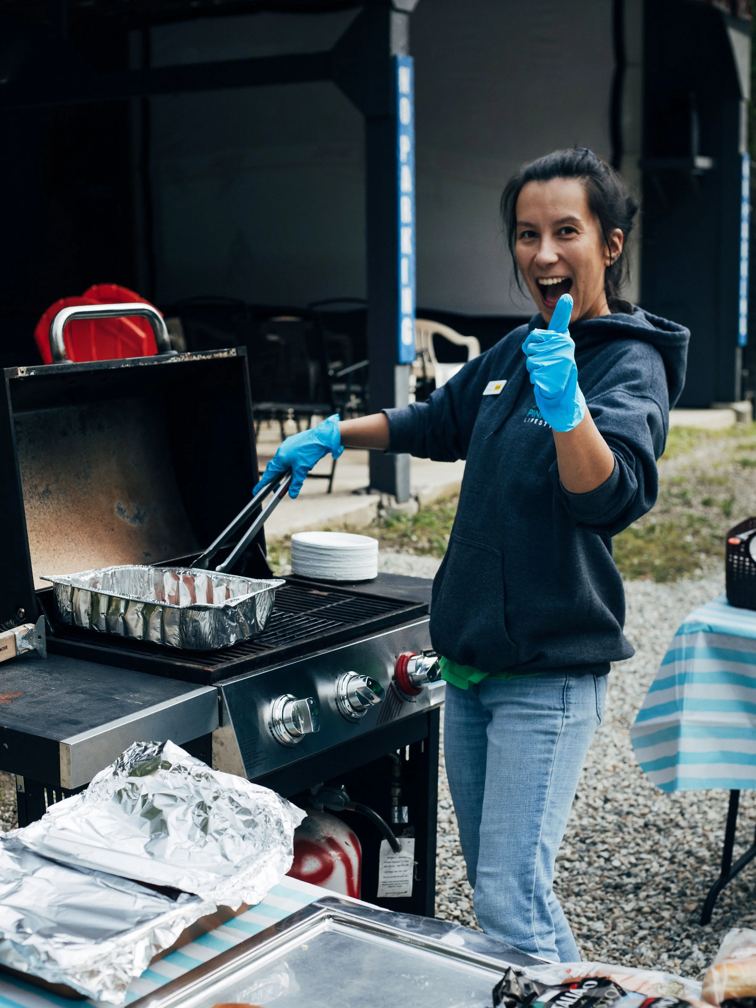 A woman wearing blue gloves and a dark hoodie smiles and gives a thumbs up while cooking on a barbecue grill outside, with a table and other items nearby.