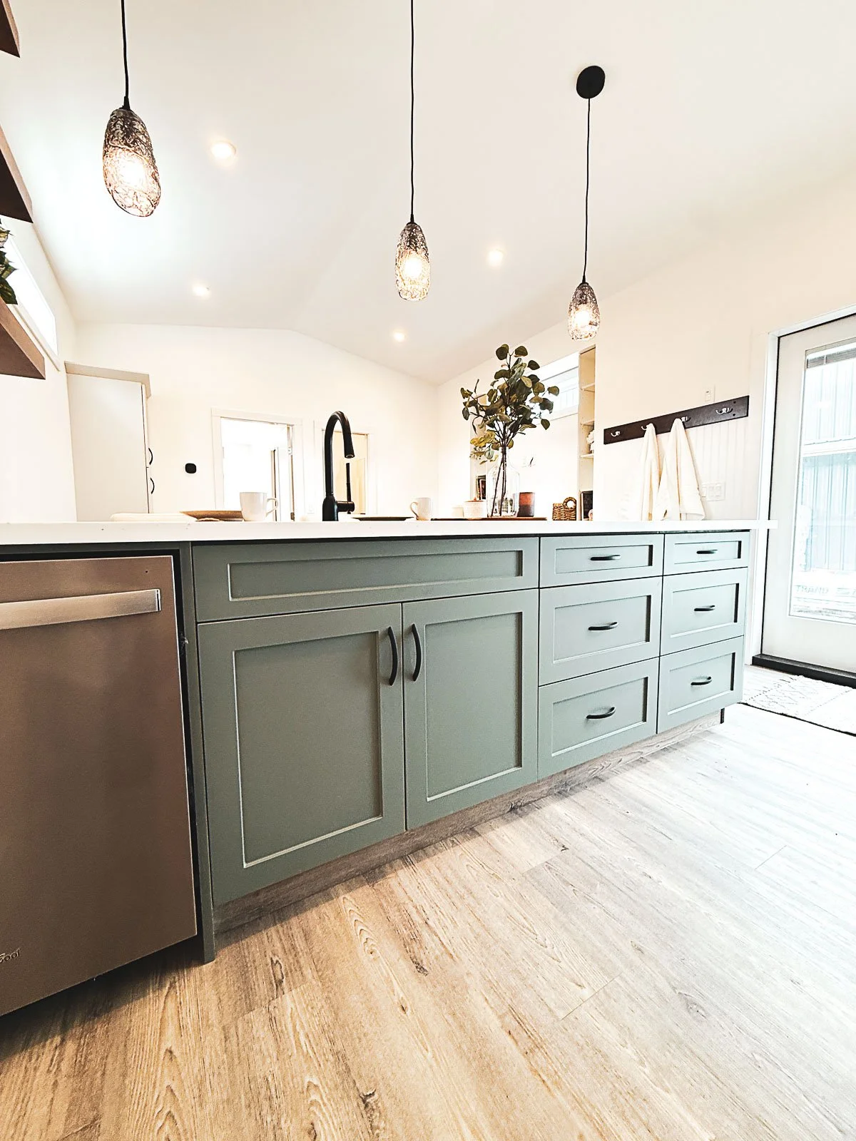 A modern kitchen with a green kitchen island featuring multiple drawers and cabinets, a black faucet, hanging pendant lights, a coffee maker, potted plant, and a sliding glass door in the background.