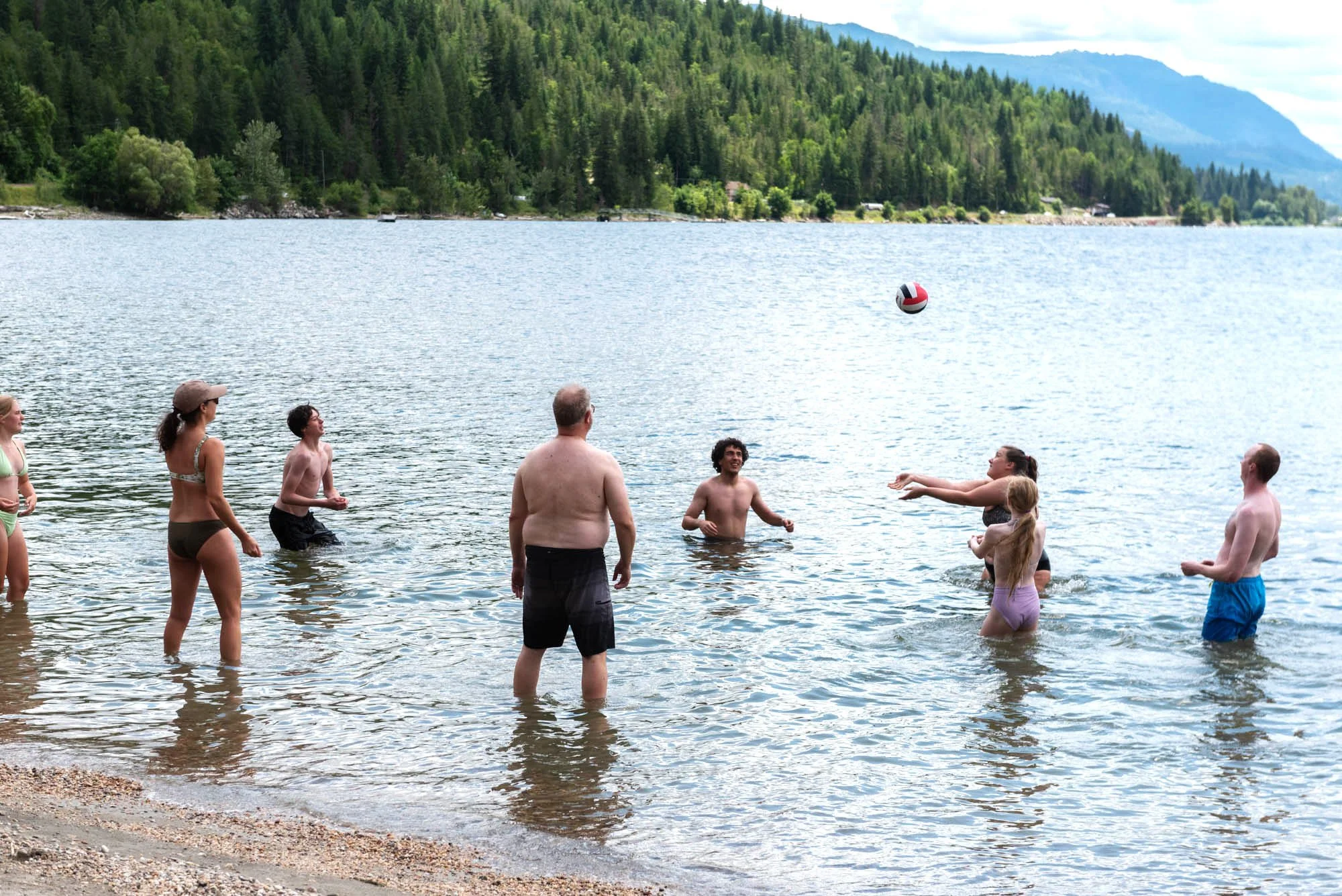 A group of people in swimsuits playing beach volleyball in a lake with mountains and forest in the background.