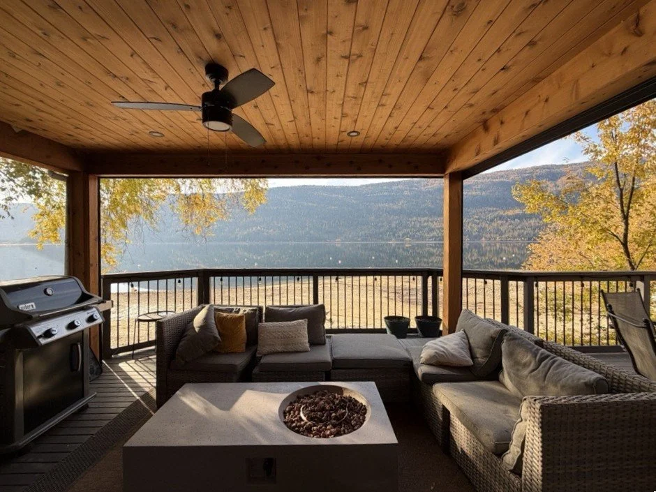 Covered outdoor porch with wooden ceiling, ceiling fan, wicker sofa with cushions, a white fire pit table, and a black grill, overlooking mountains and trees.