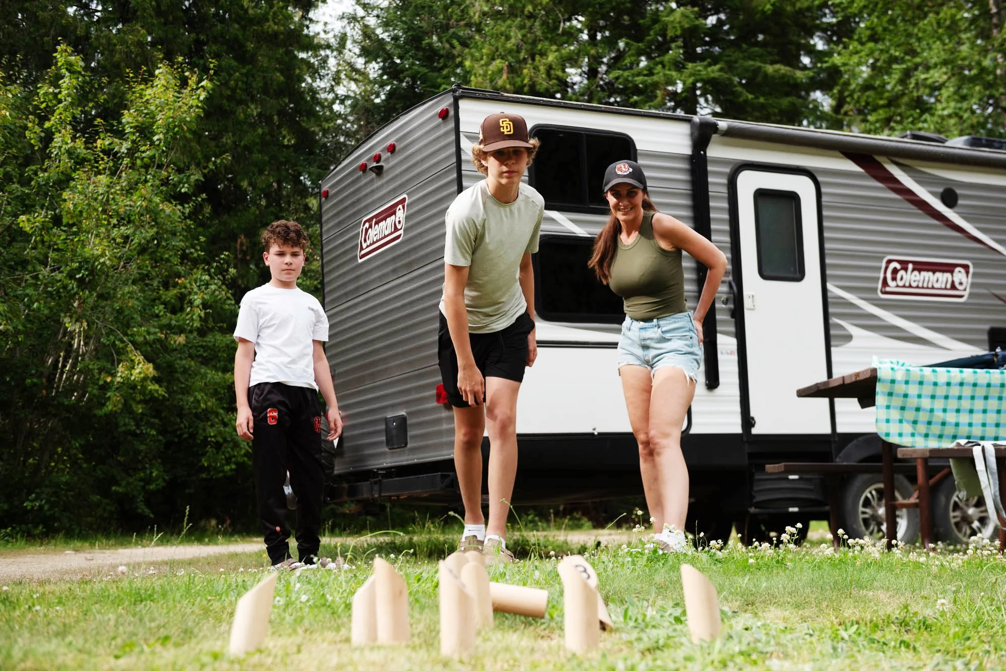 Three children outdoors near a camper trailer, playing a game with wooden pins set up on the grass.