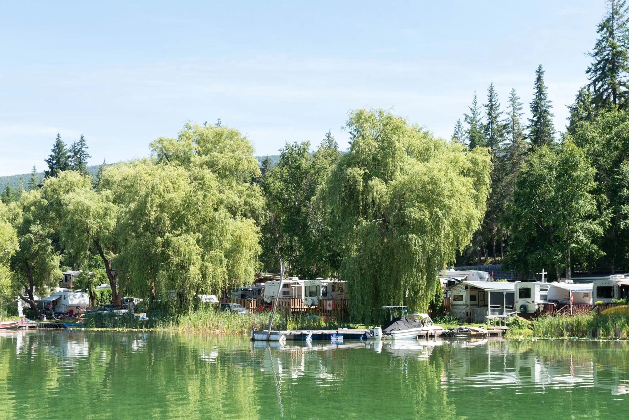 A lakeside camping area with trees, RVs, and boats on the water, under a blue sky.