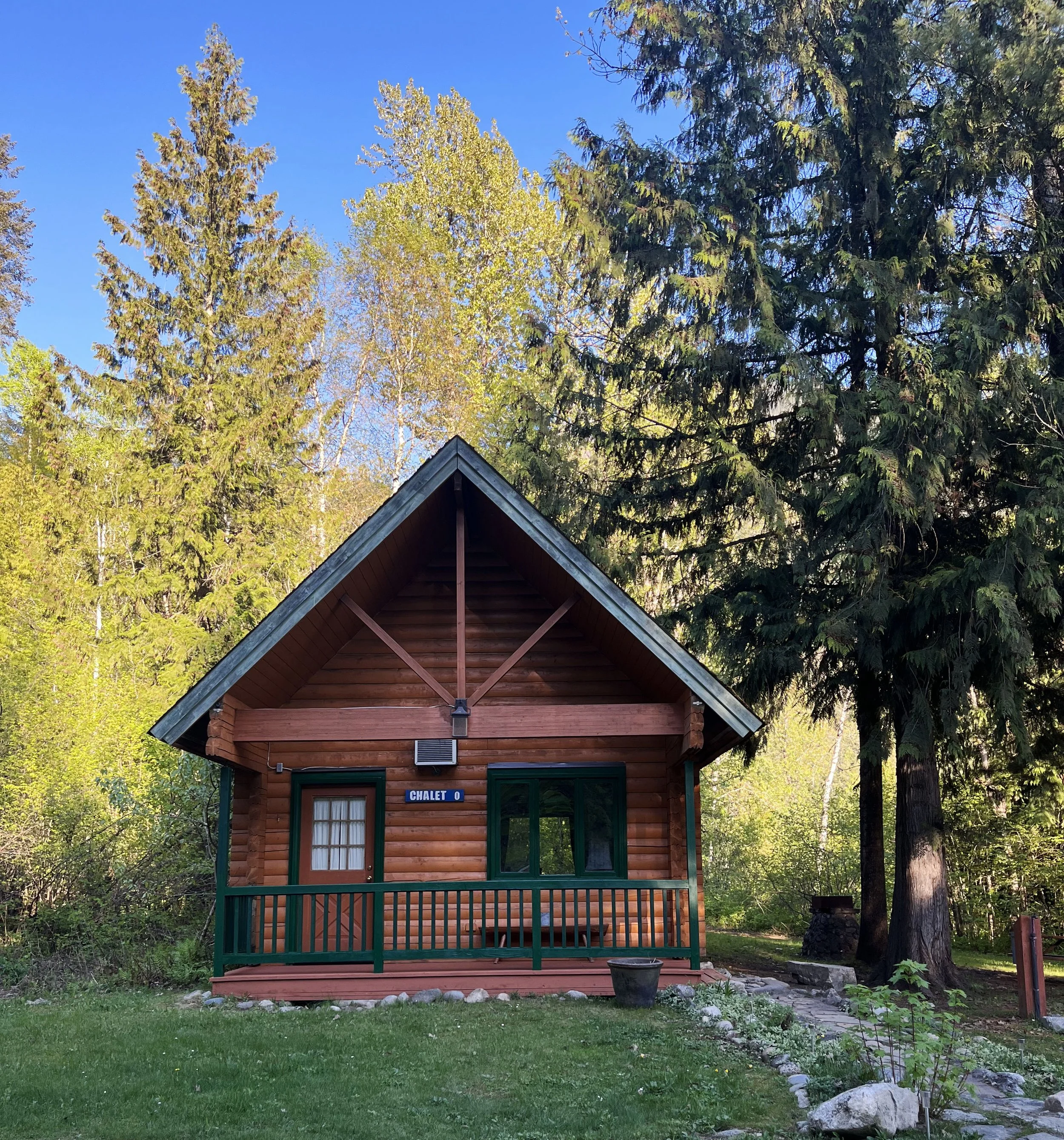 A small wooden chalet with a dark gray roof, surrounded by green grass and trees, with a garden bed along the right side and a small gravel pathway leading to the front porch. The chalet has a sign that reads 'CHALET 0'.