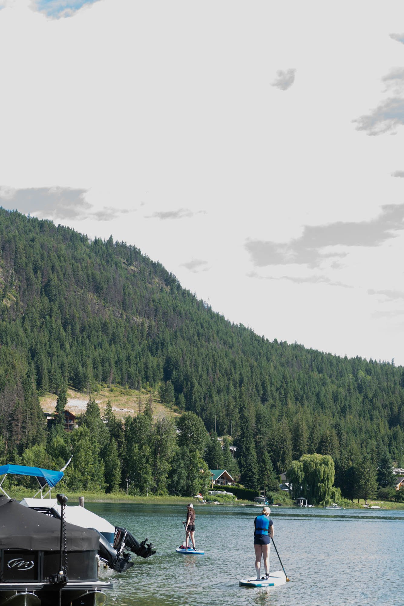 People paddleboarding and standing in a lake with boats docked at the shore, surrounded by green forested hills with houses.
