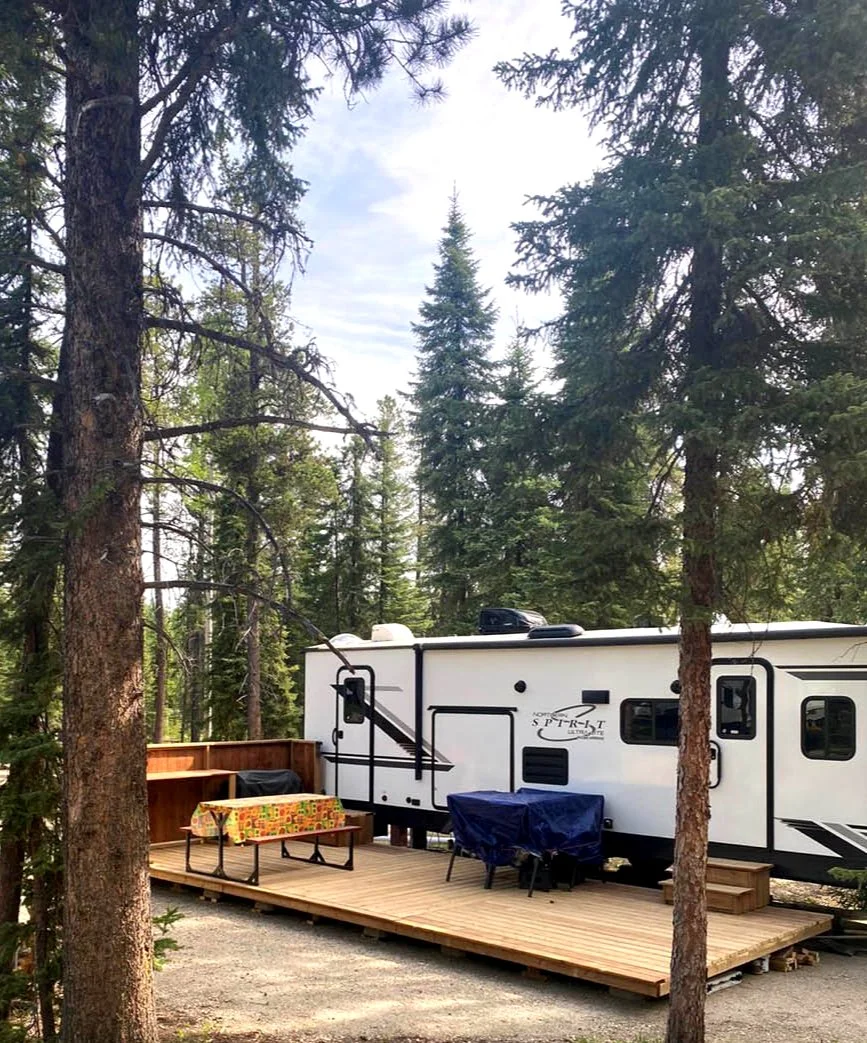 A travel trailer is parked on a wooden deck in a forested camping area, with tall pine trees surrounding it and a partly cloudy sky overhead.