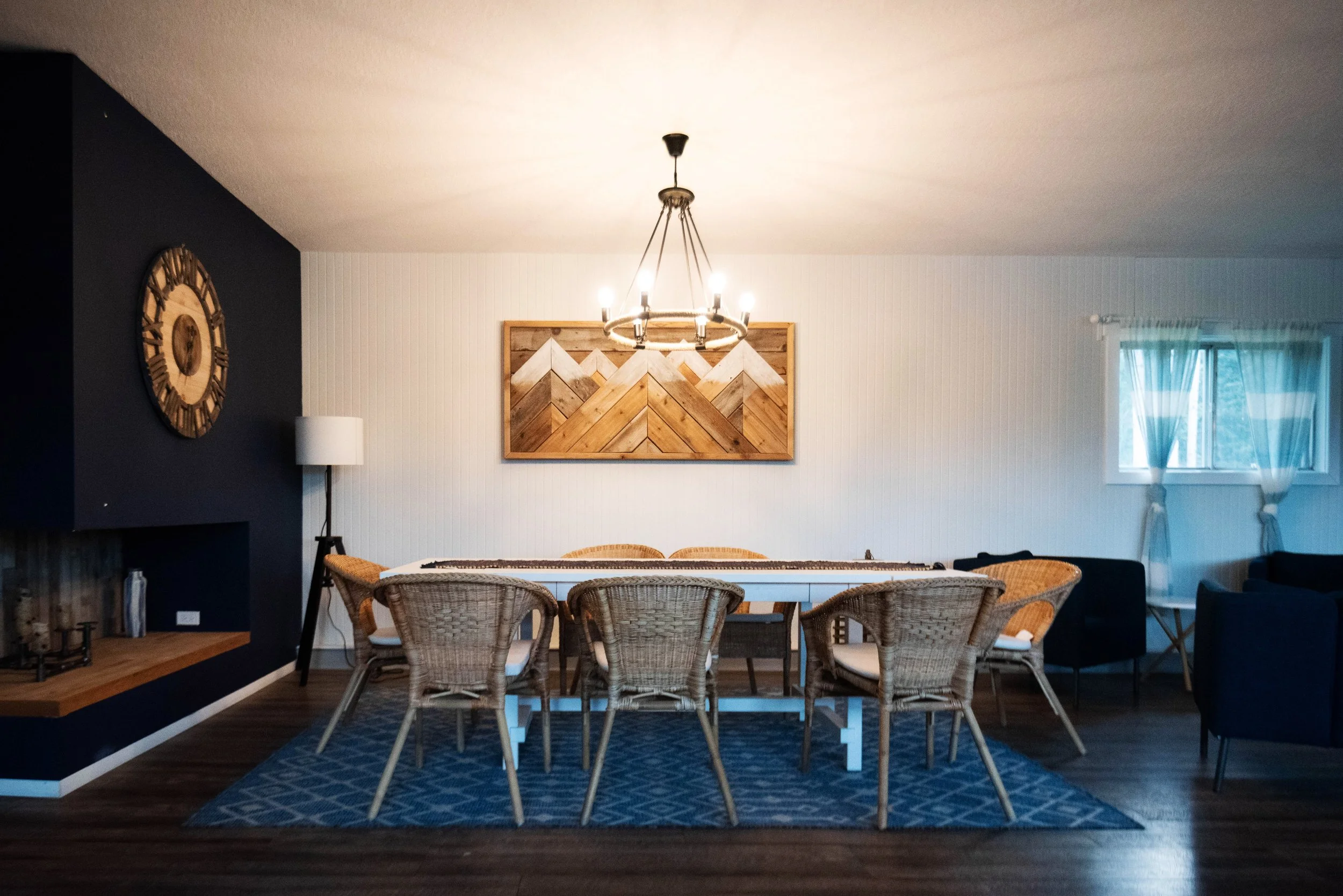 A dining room with a white table surrounded by wicker chairs on a blue patterned rug. A wooden wall art piece and a large clock are on the white and dark walls. A chandelier hangs above the table, and a window with sheer curtains lets in natural ligh